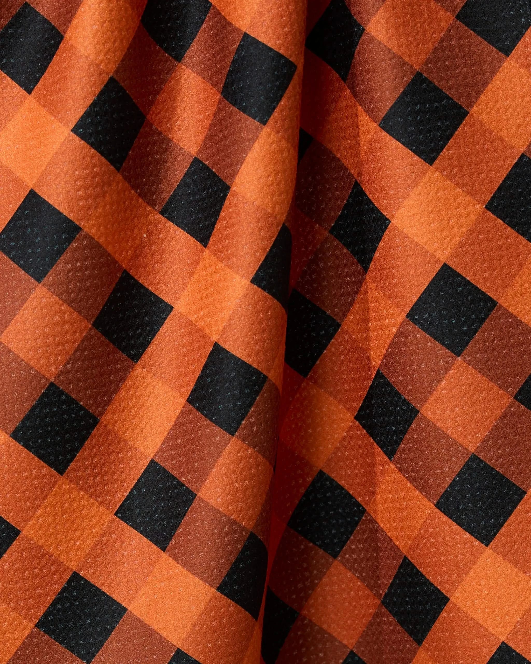 A close-up of Geometrys Pumpkin Plaid tea towel shows its orange and black checkered pattern, textured surface, and soft folds, all crafted from recycled materials.