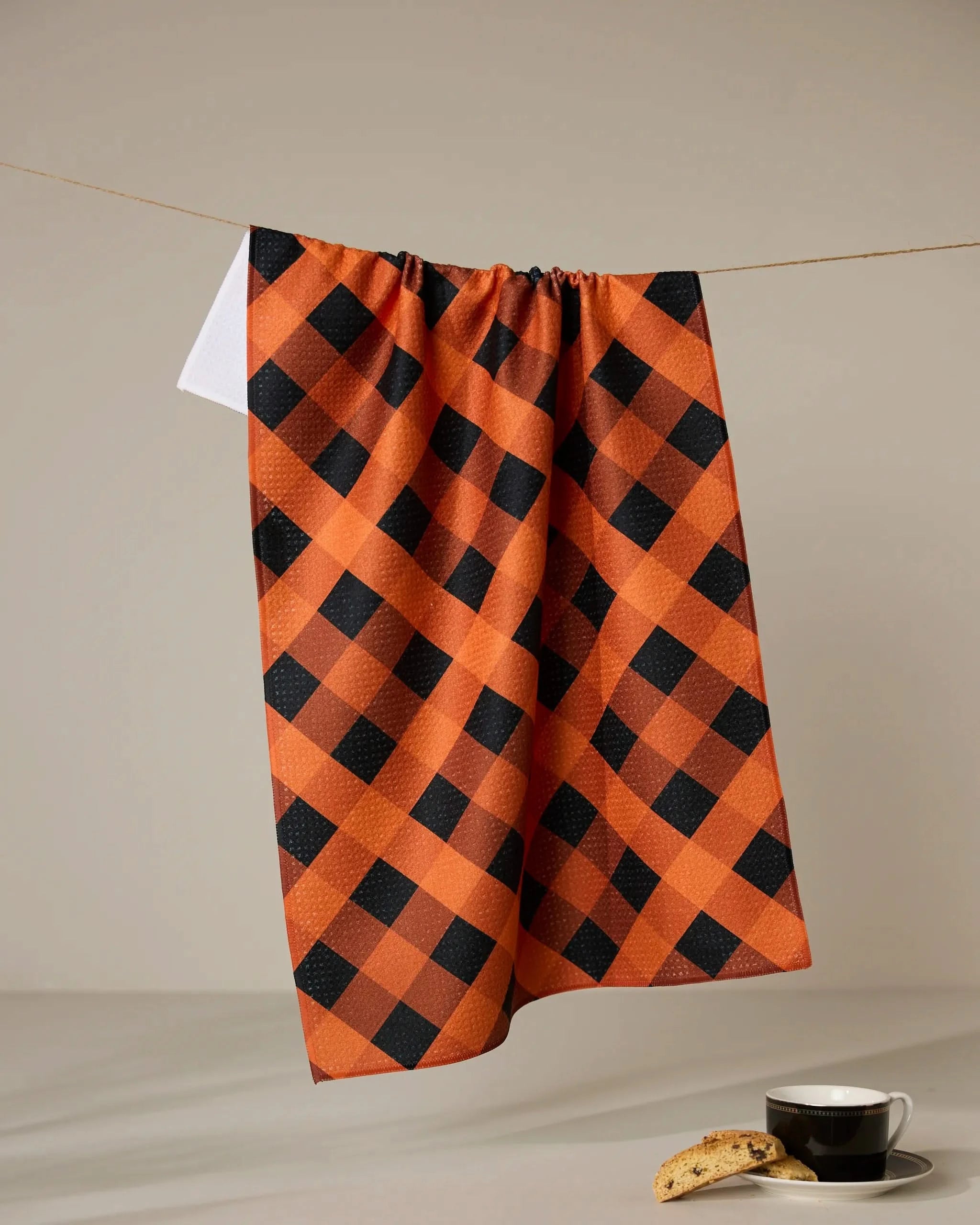 The Pumpkin Plaid tea towel by Geometry, crafted from recycled materials in an orange and black checkered pattern, hangs on a string above a black coffee cup and biscotti on a light surface with a clean, neutral background.