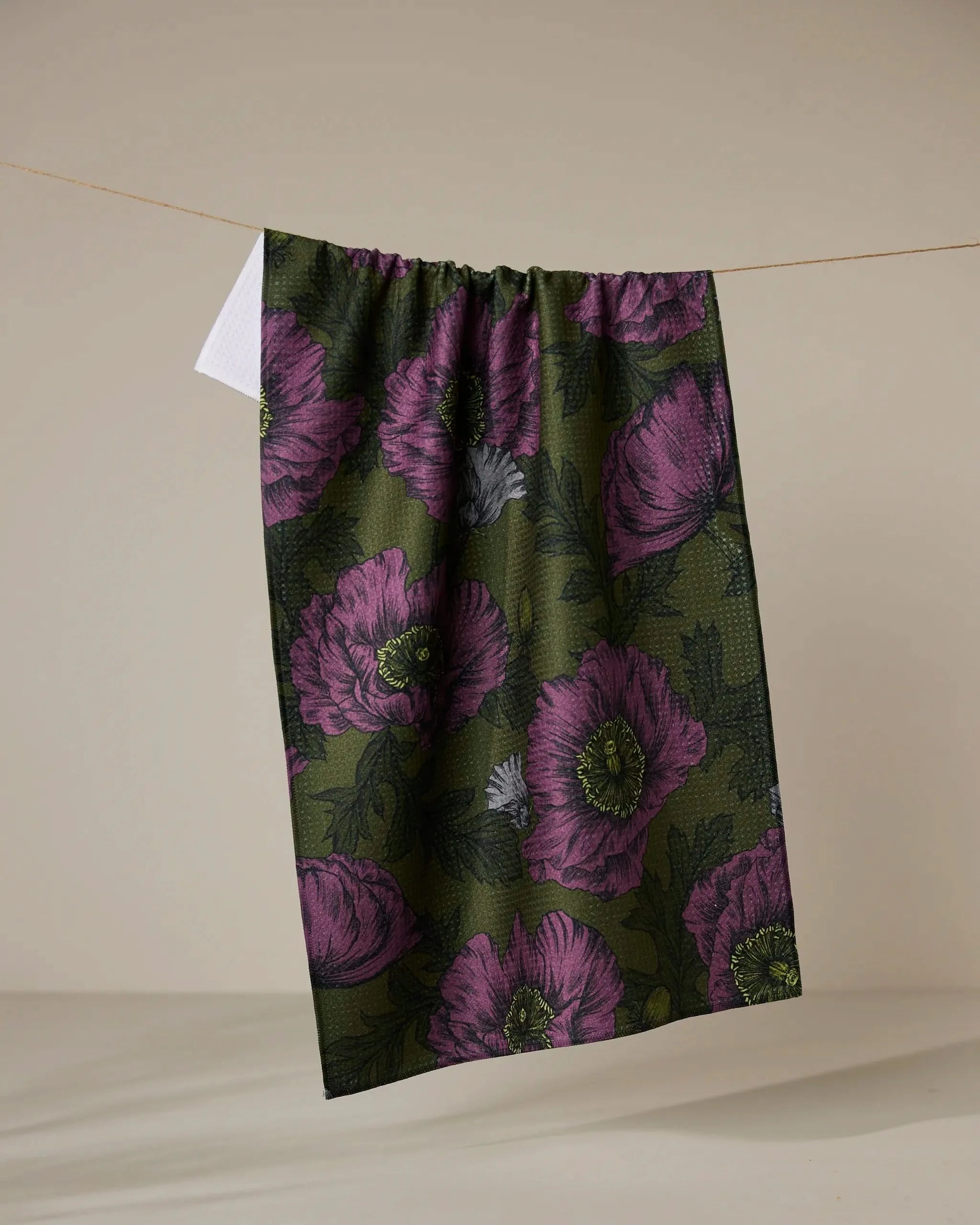 Geometry’s Potion Poppies tea towel, made from recycled materials, features large purple and light gray floral patterns on dark green and is shown hanging against a beige background.