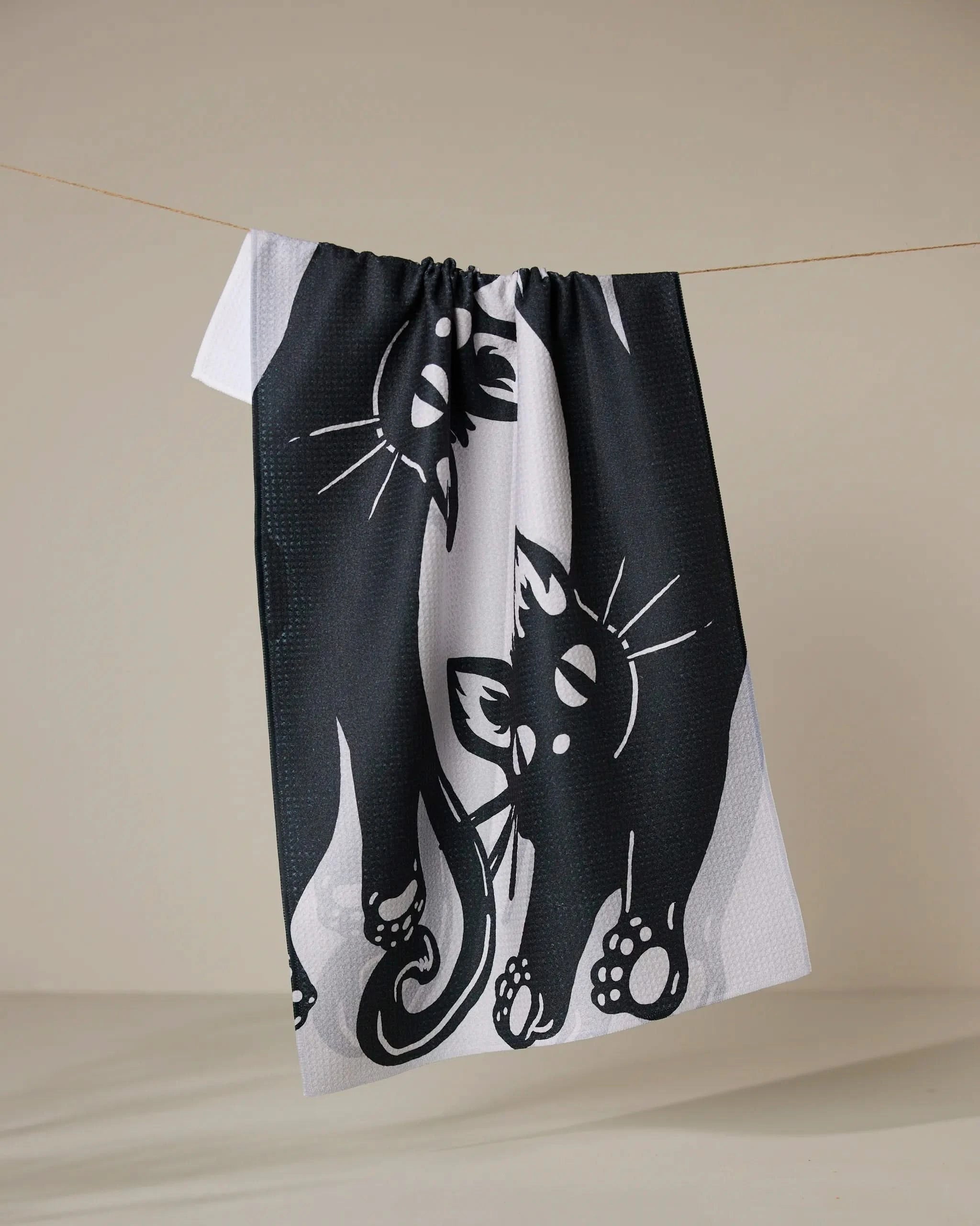 The Geometry Night Zoomies tea towel, crafted from recycled materials, showcases a playful black and white cat illustration and hangs on a string against a plain beige background.