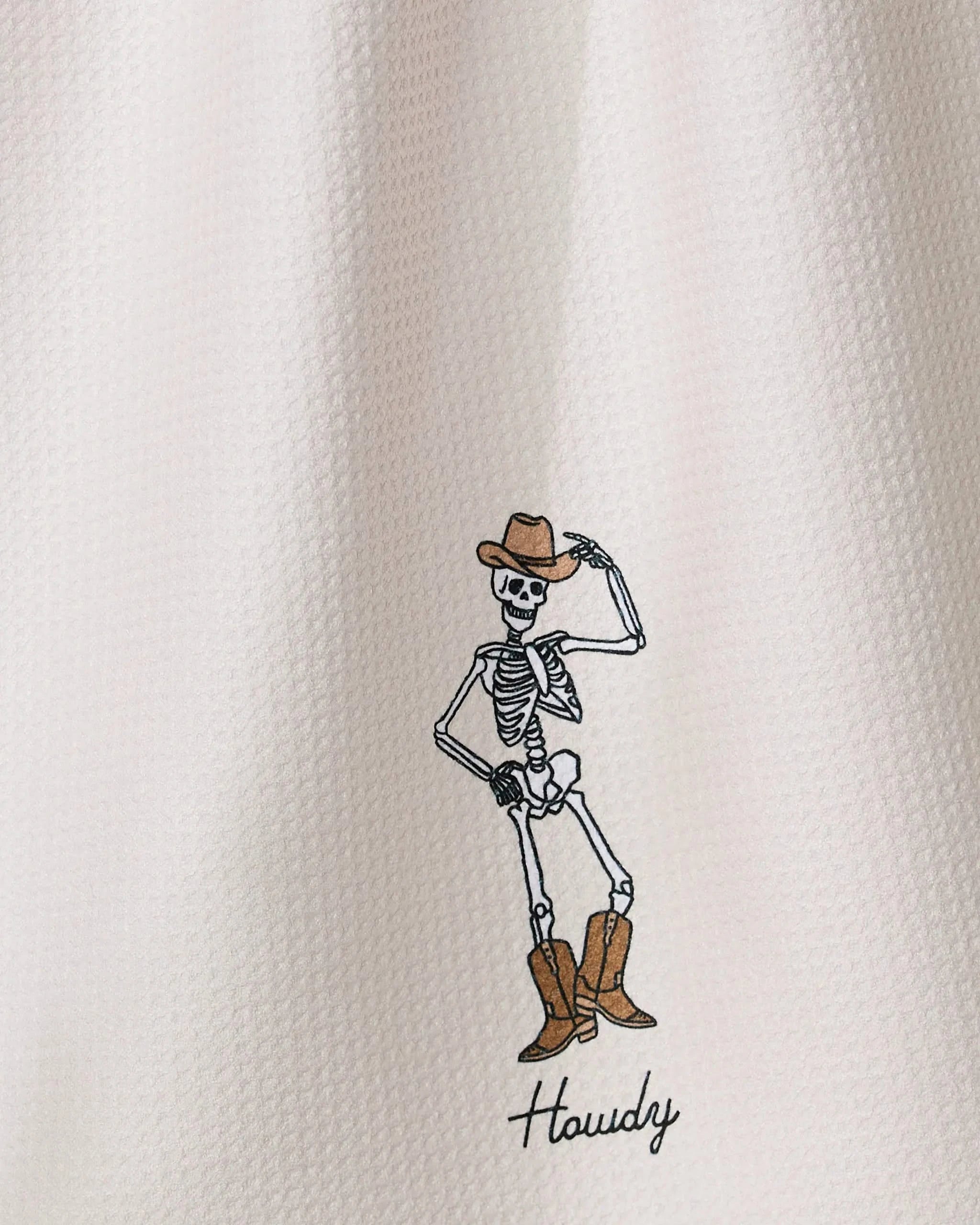 The Geometry Howdy Cassondra features a cartoon skeleton in a cowboy hat and boots, striking a pose and tipping its hat, with Howdy below on a quick-drying tea towel made from recycled materials and a white textured background.