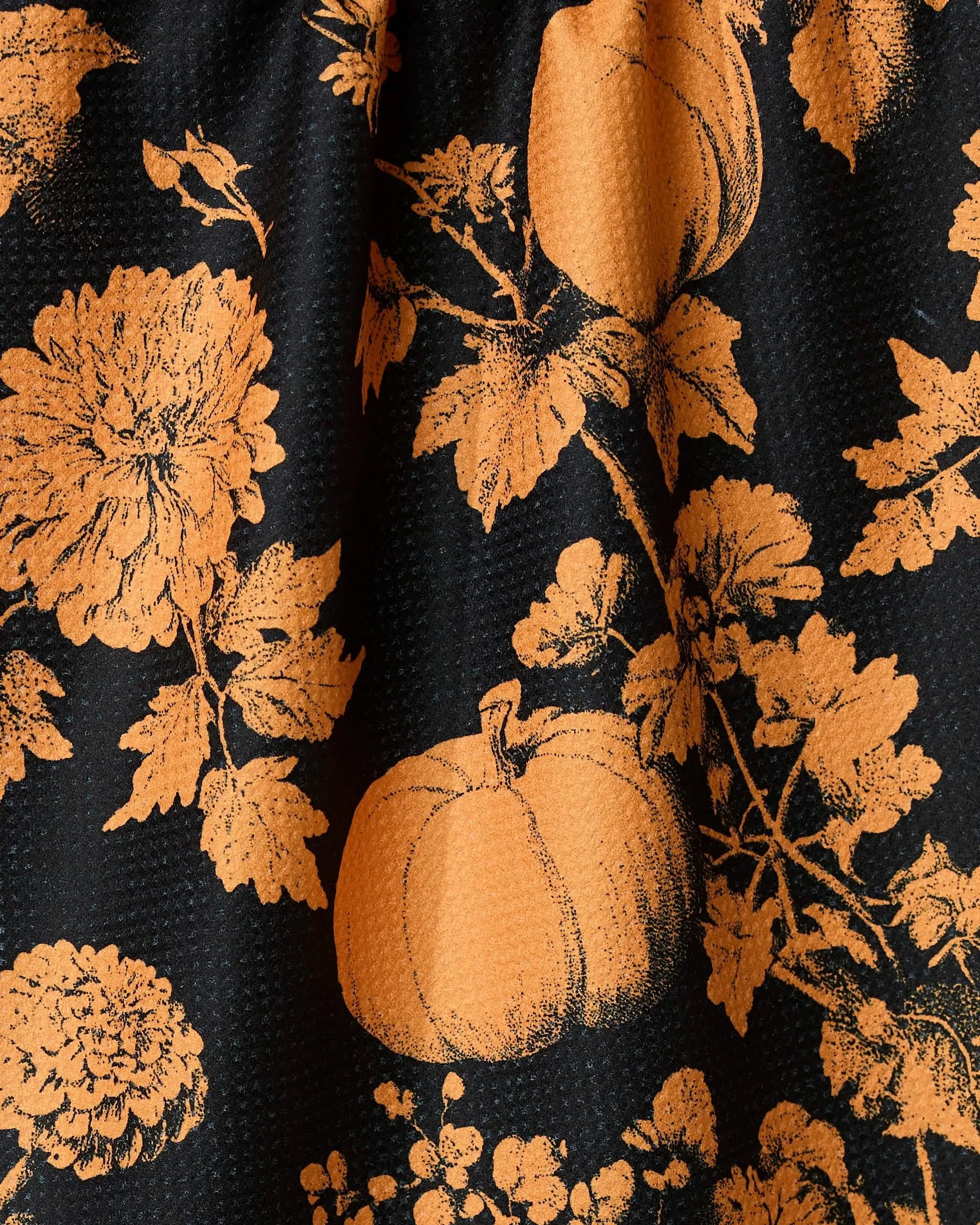 The Haunted Harvest Orange tea towel by Geometry features orange pumpkins and flowers on black textured fabric, creating a festive autumnal pattern. This quick-drying towel is perfect for seasonal decor.