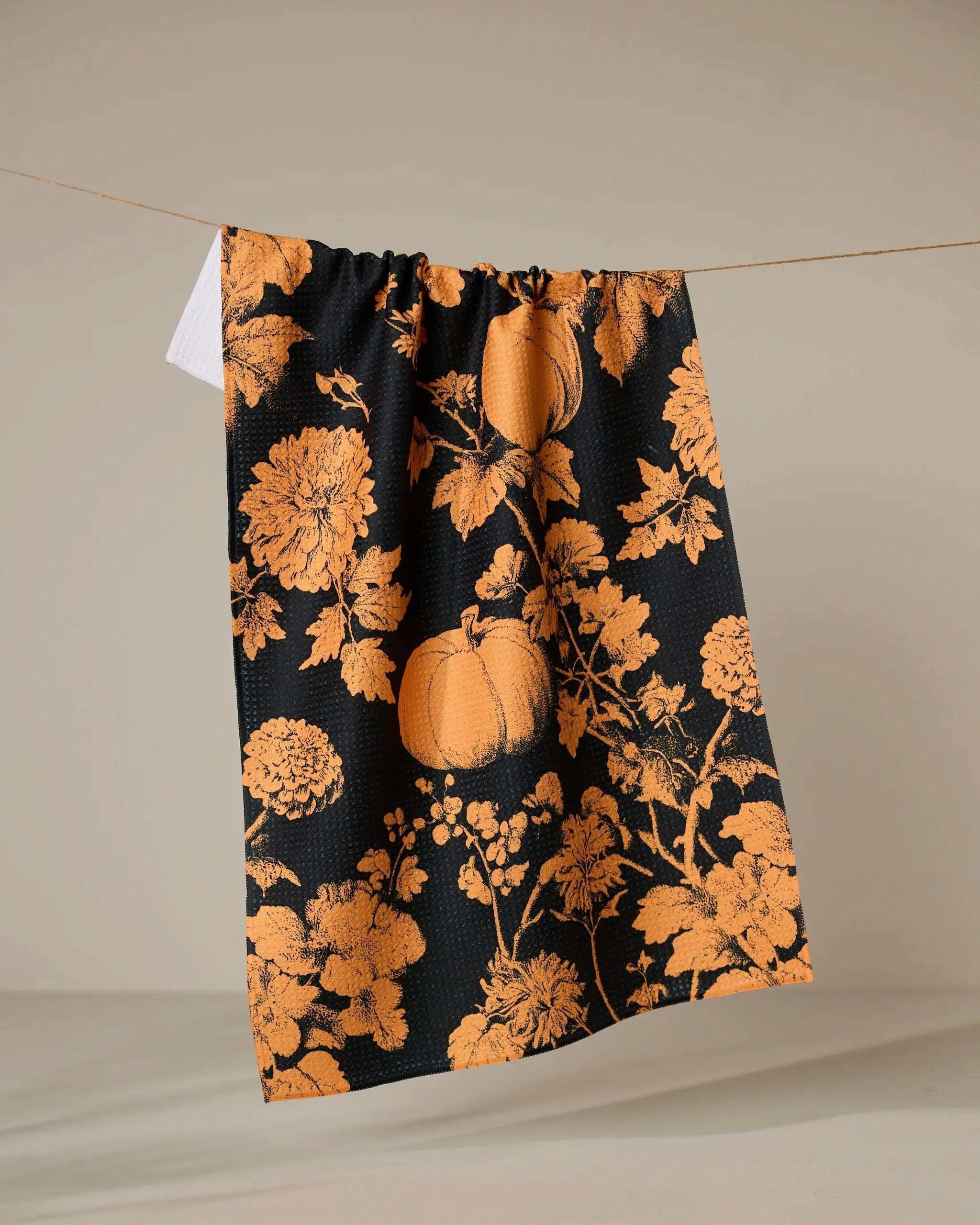 The Geometry Haunted Harvest Orange recycled kitchen towel, featuring an orange floral and pumpkin pattern on black, hangs on a line against a plain, light background.