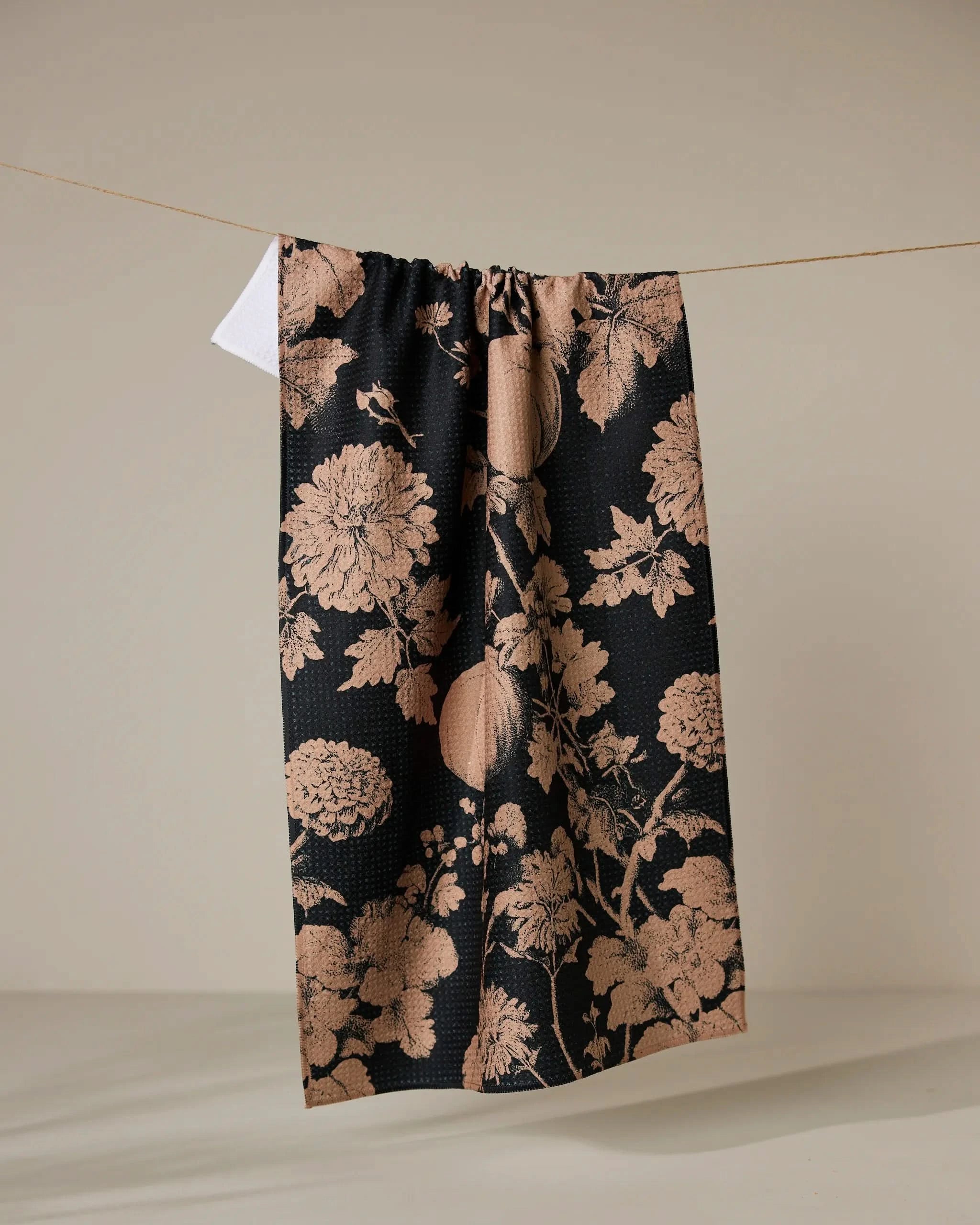 The Geometry Haunted Harvest black tea towel, featuring a peach floral pattern and made from recycled materials, hangs on a thin clothesline against a plain beige background.