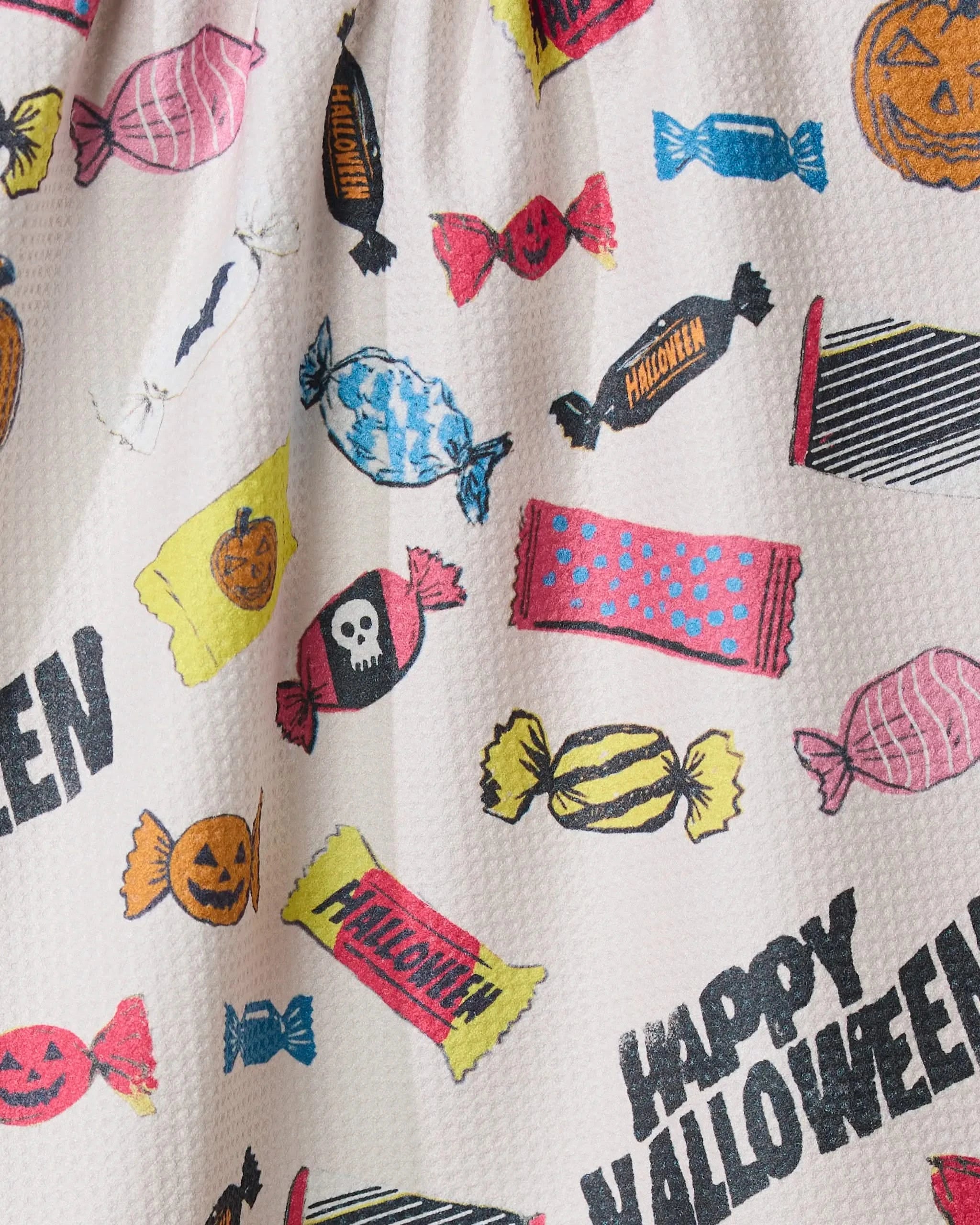 The Geometry Halloween Treats tea towel features vibrant Halloween candy, pumpkin, and skull illustrations with Happy Halloween text, printed on textured white fabric made from recycled materials.
