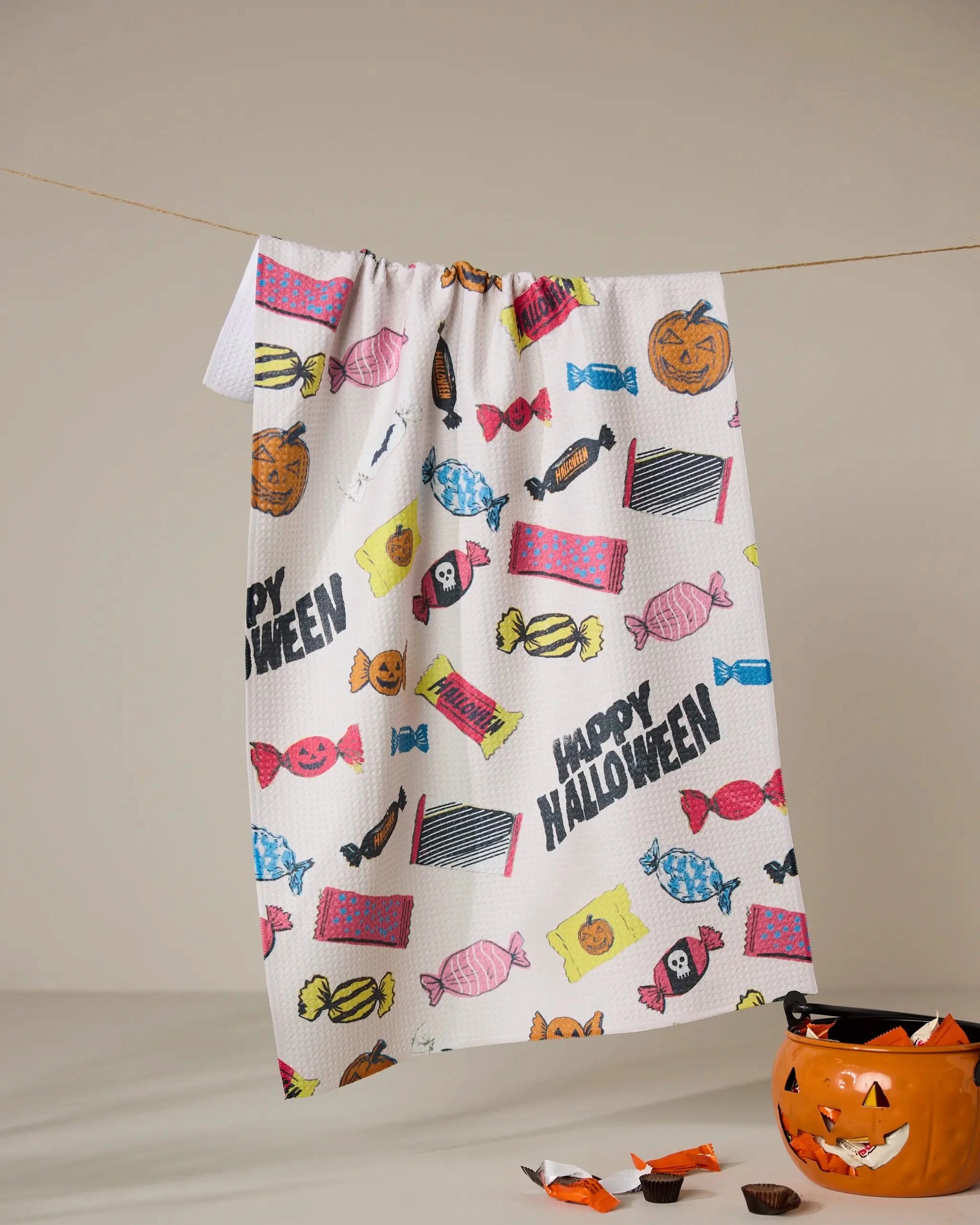 The Geometry Halloween Treats towel is made from recycled materials and showcases vibrant Halloween-themed designs—candies, jack-o’-lanterns, black cats, and festive text—with a pumpkin-shaped candy container adding spooky cheer.