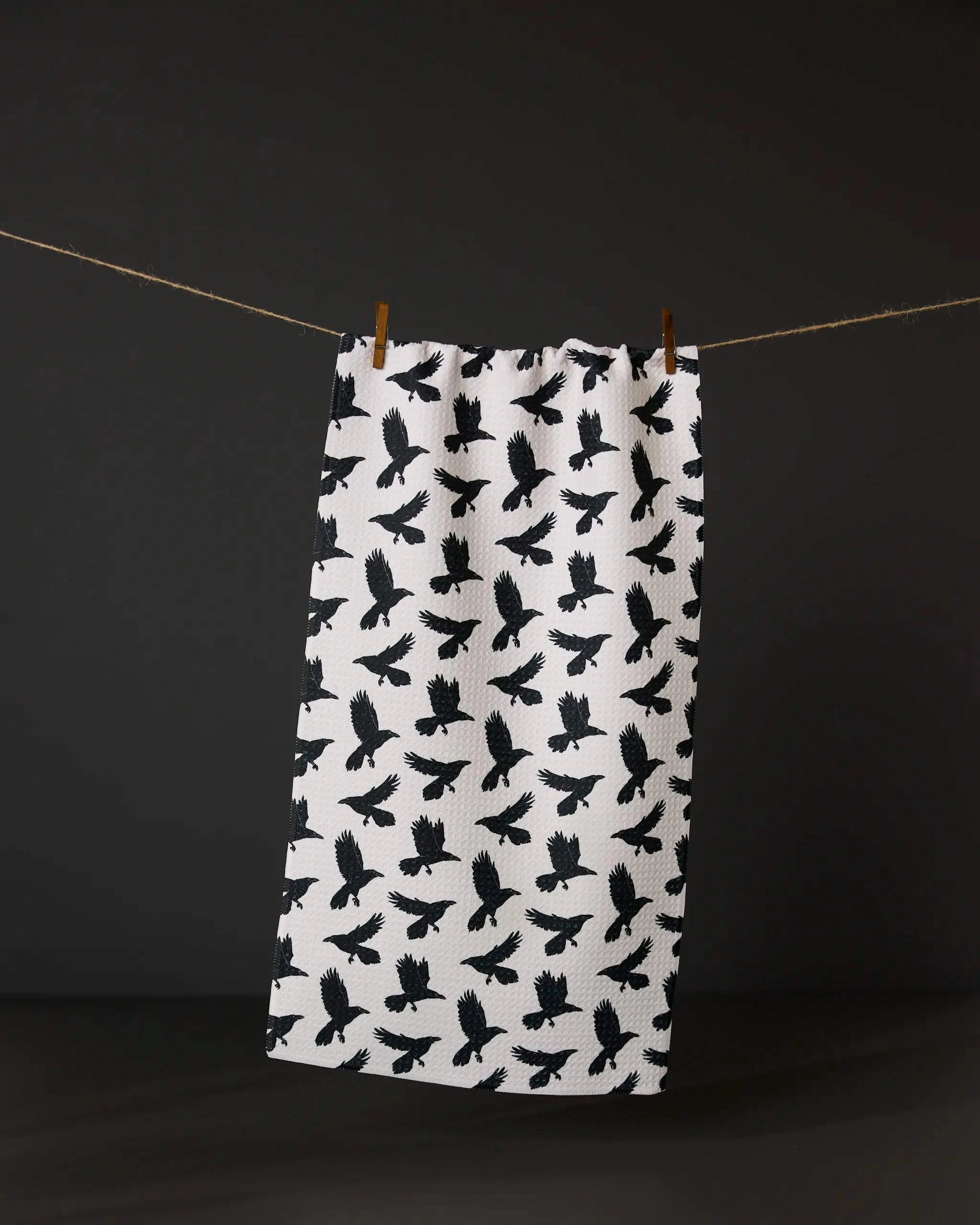 Crow Call by Geometry: a white bar towel featuring a repeating black crow pattern hangs on a clothesline with two wooden clothespins, set against a dark background.