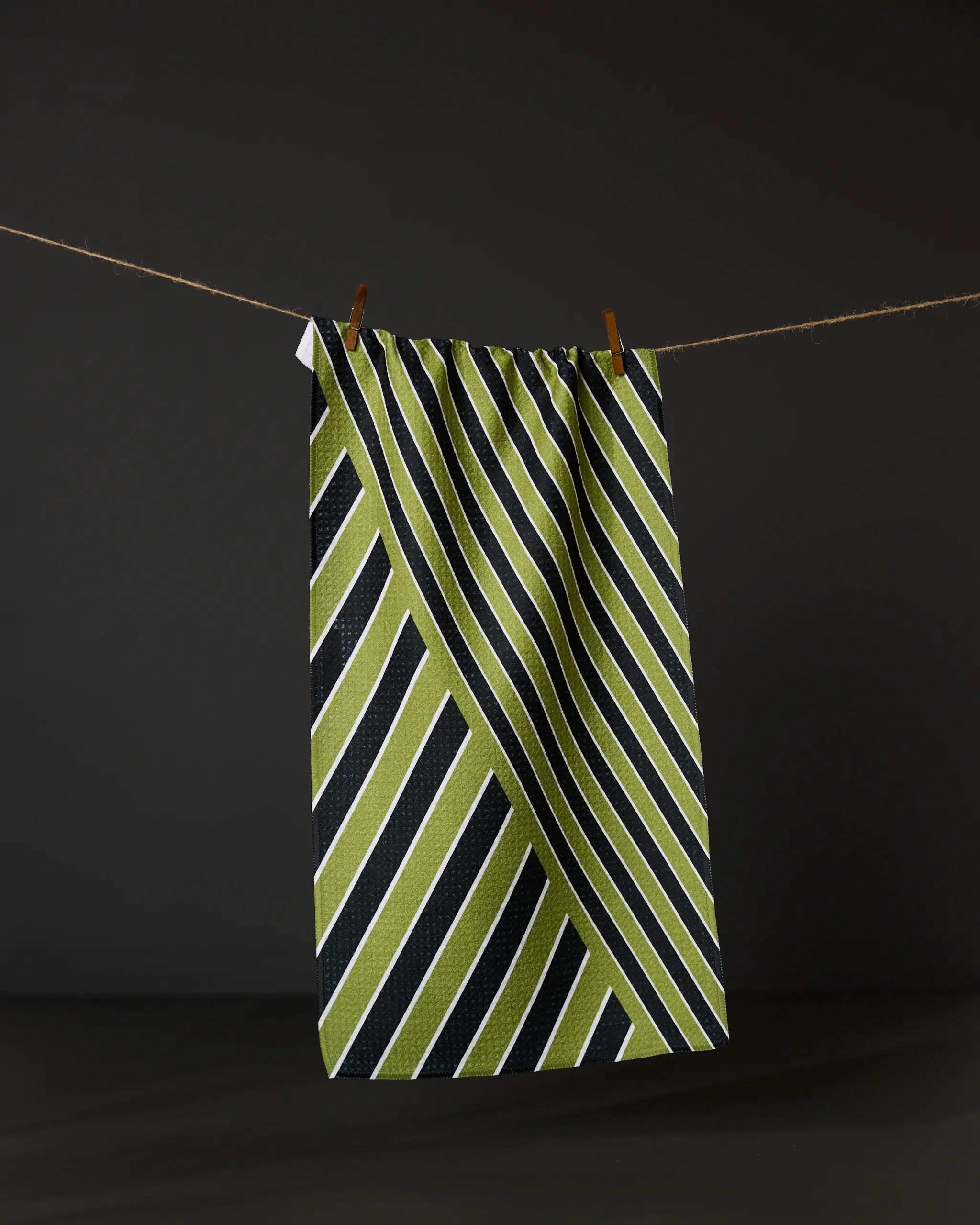 The Geometry Tricky Lines towel, featuring green, black, and white diagonal stripes, hangs on a clothesline with two wooden clothespins against a dark background.