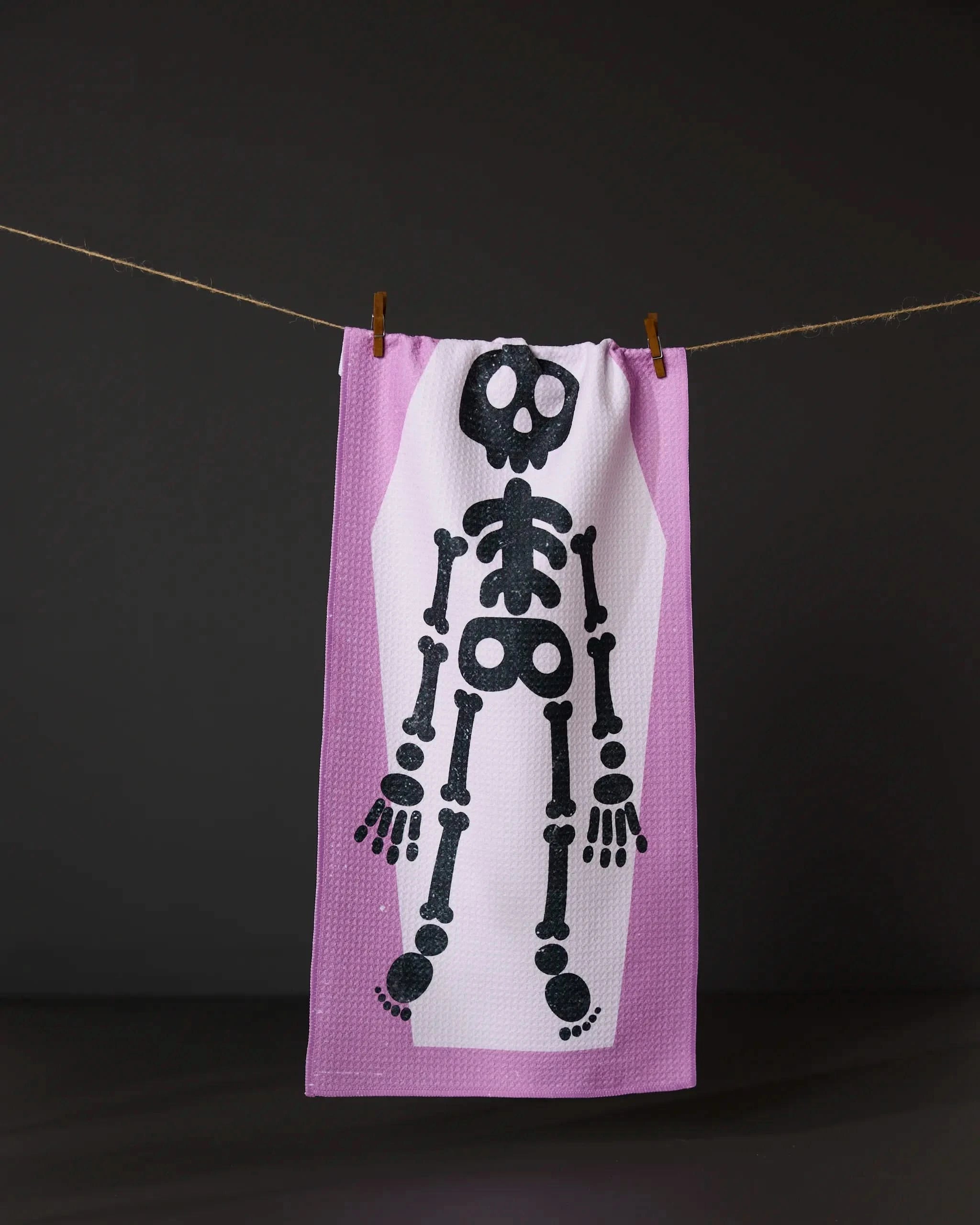 The Bright Bones towel by Geometry is ultra-absorbent, white with a purple border, and features a cartoon skeleton design, shown hanging on a clothesline against a dark background.