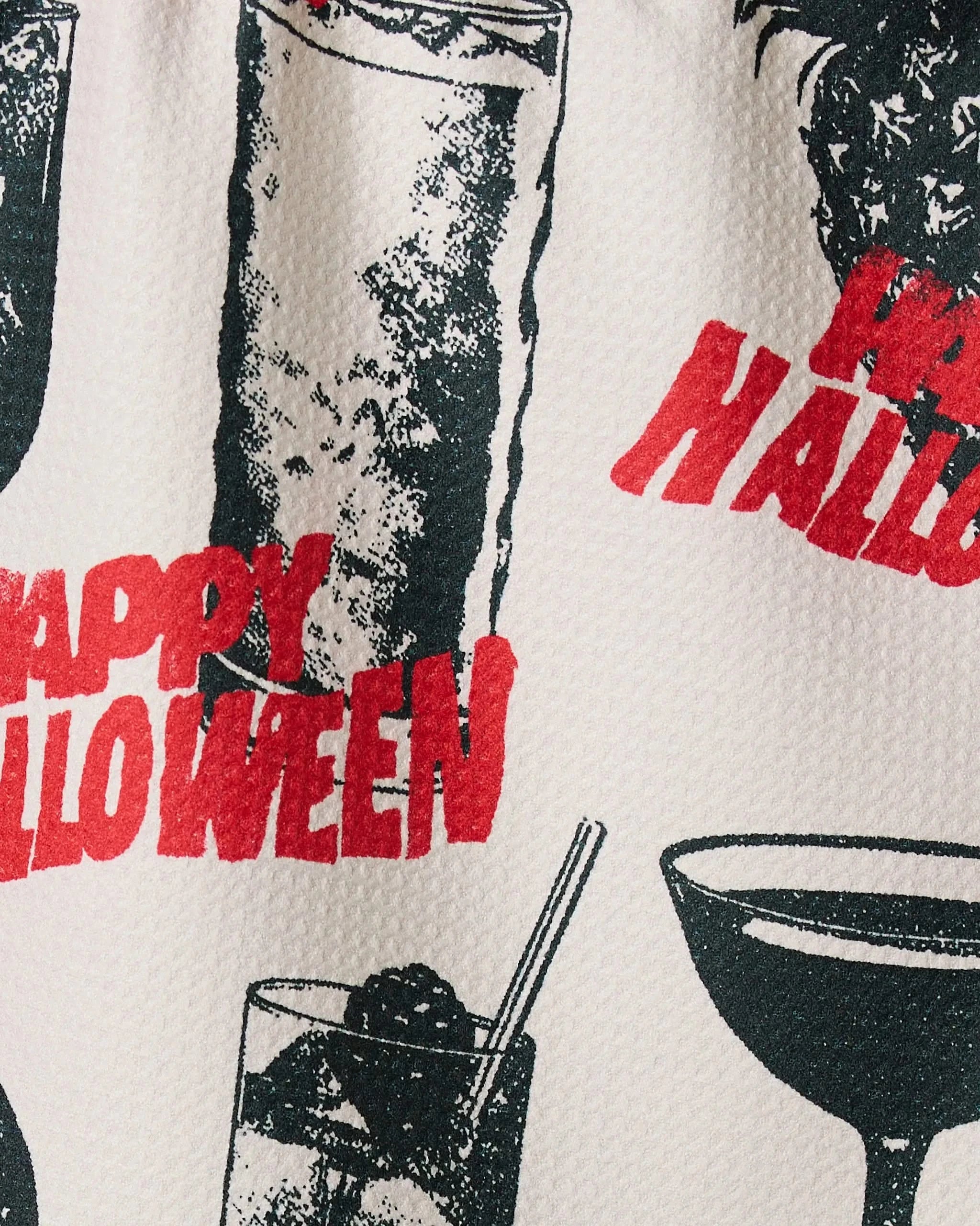 Close-up of Geometrys Ghostly Cheers bar towel featuring Halloween-themed cocktail prints and the phrase Happy Halloween in bold red on a textured white background.