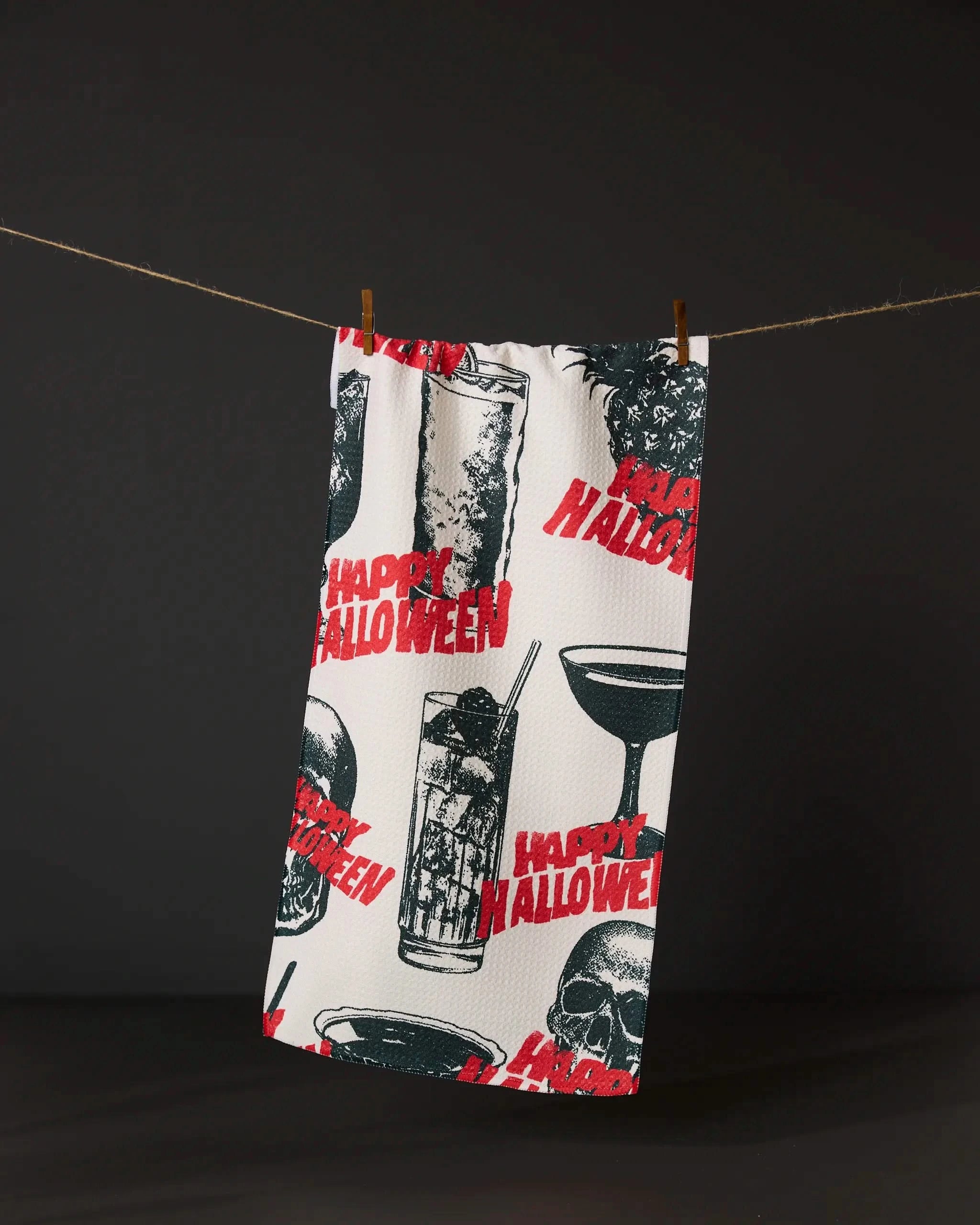 The Geometry Ghostly Cheers bar towel displays black and white skulls, cocktails, and bold red Happy Halloween text, shown hanging on a clothesline with a dark backdrop.
