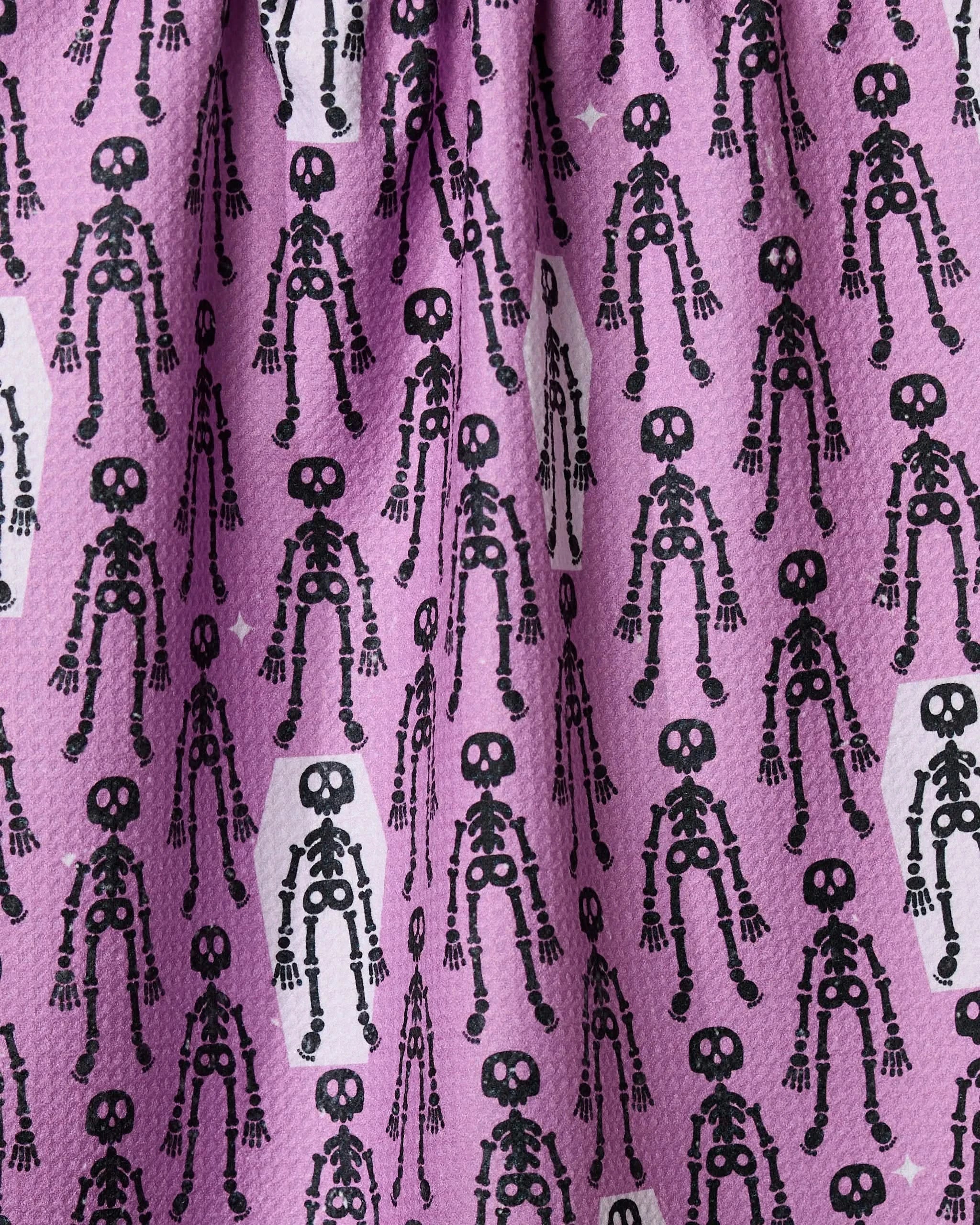 A close-up of Geometry’s “Resting Skeletons” tea towel showcases pink fabric with playful black cartoon skeletons, adding whimsical charm and functional style to any kitchen.