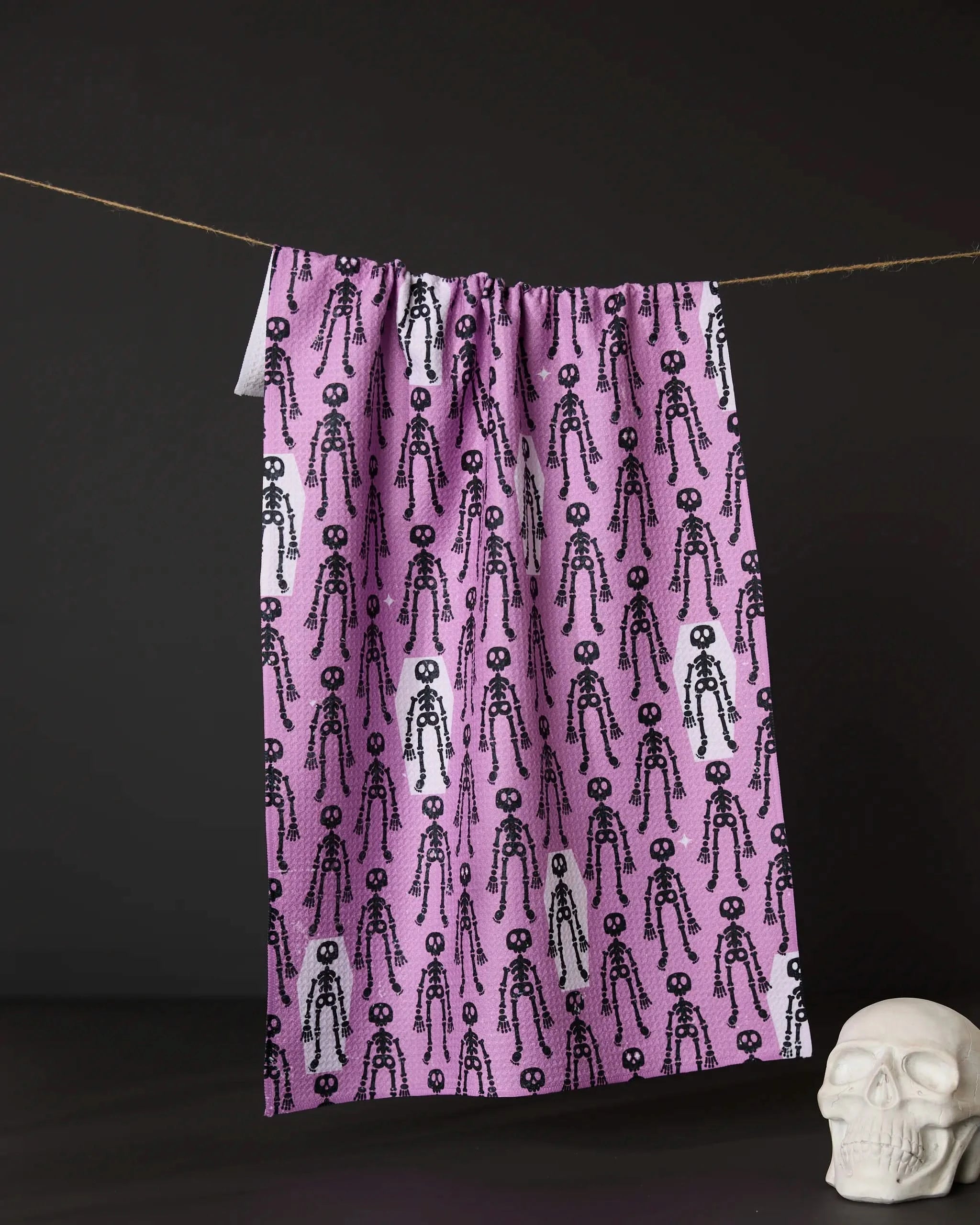 The Geometry Resting Skeletons pink recycled kitchen towel, featuring black and white cartoon skeletons, hangs from a string against a dark background with a white skull decor resting nearby.