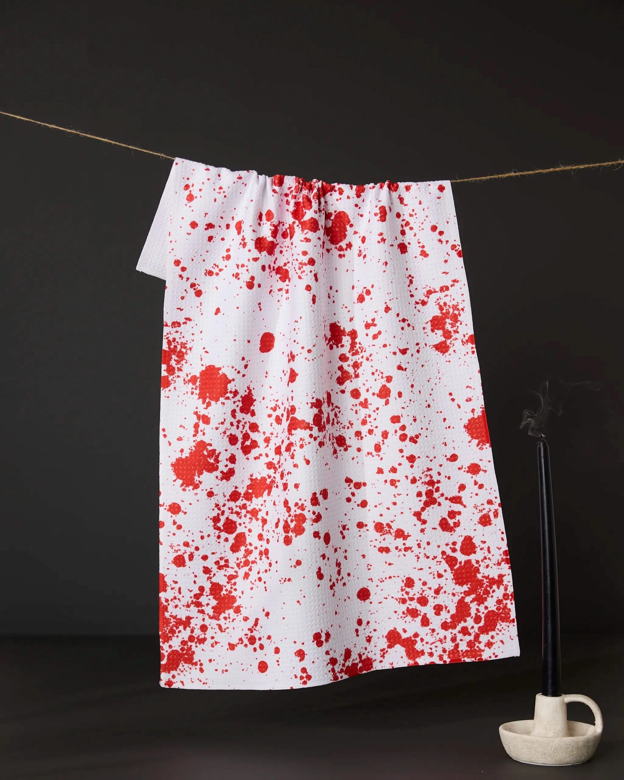 The Geometry Zombie Brains kitchen towel, featuring eco-friendly white fabric with red splatter patterns, hangs beside a thin black candle in a holder, its gentle trail of smoke set against a dark backdrop.