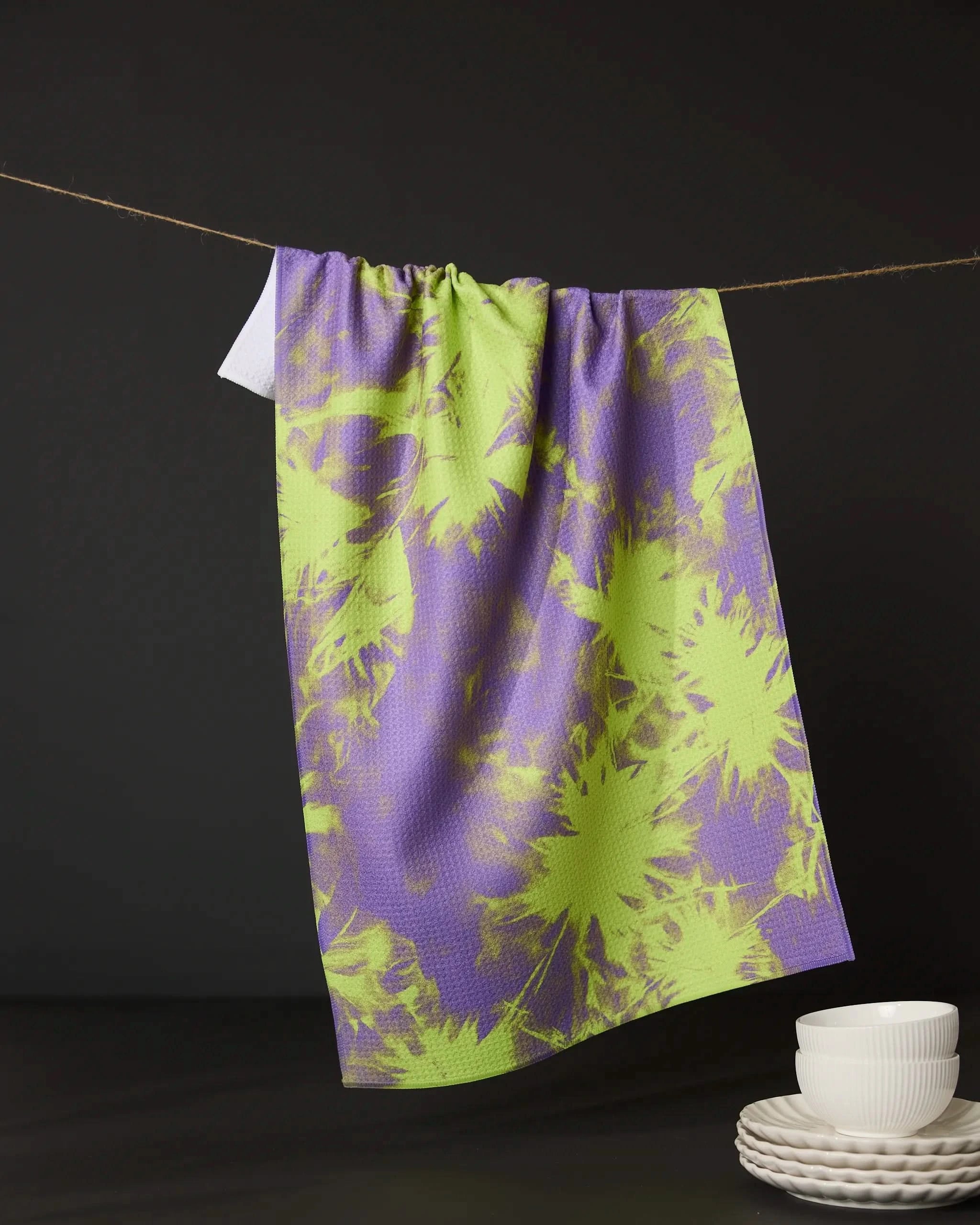 Geometry’s Potion Purple tea towel, featuring a purple and green tie-dye design and made from recycled materials, hangs on a clothesline against a black background, with white plates and bowls stacked below on the right.