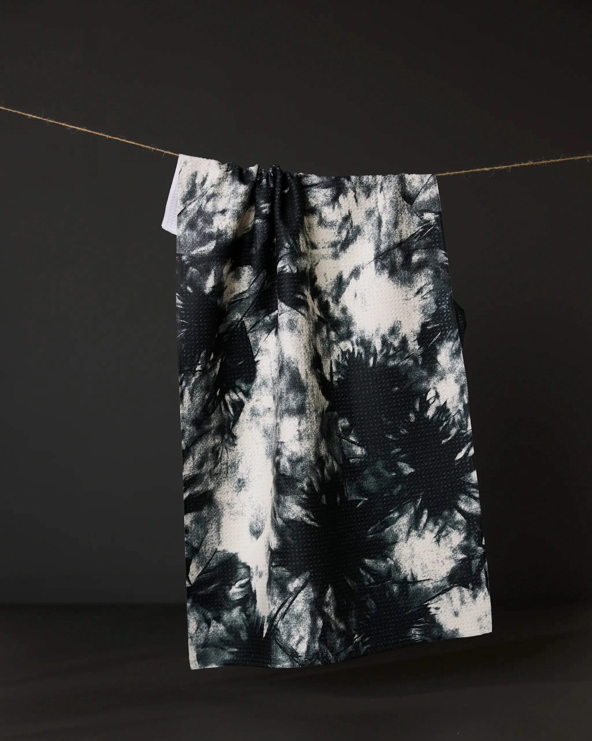 The Geometry Potion tea towel, featuring a black and white tie-dye design, hangs on a string against a dark background.