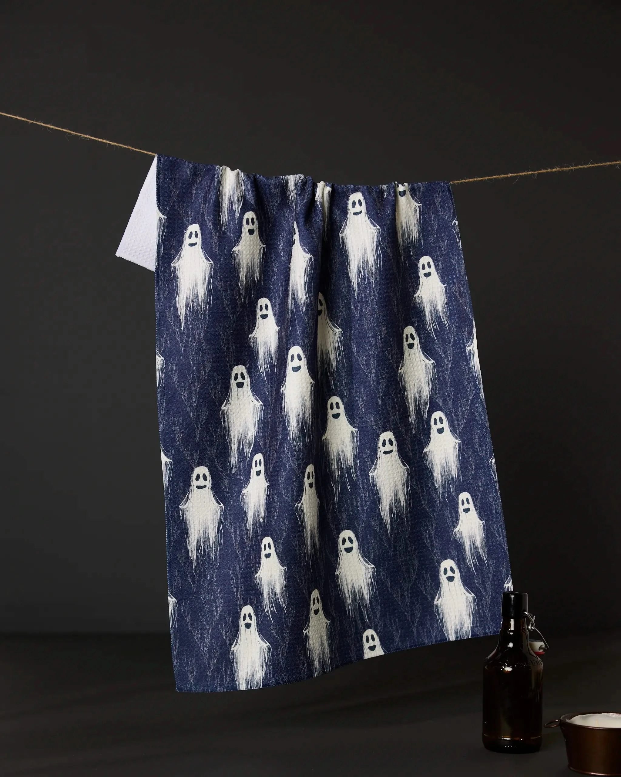 The Geometry Happy Ghosting kitchen tea towel, featuring white cartoon ghosts on dark blue, hangs on a clothesline against a dark background with a dark glass bottle and cup visible below.