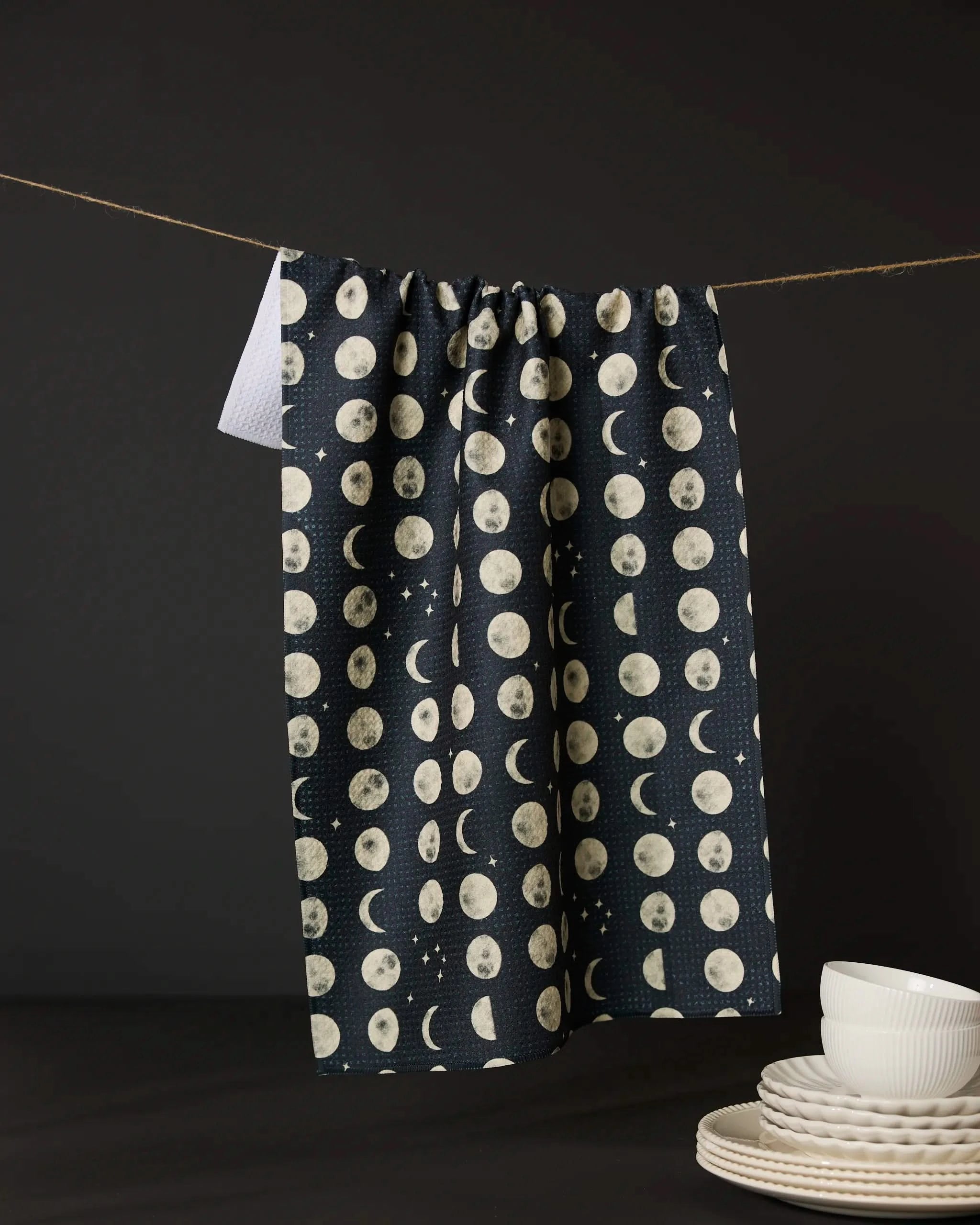 The Geometry Moon Dots tea towel, ultra-absorbent and dark with moon phase patterns, hangs on a string. Stacks of white dishes and bowls sit in the lower right corner against a black backdrop.