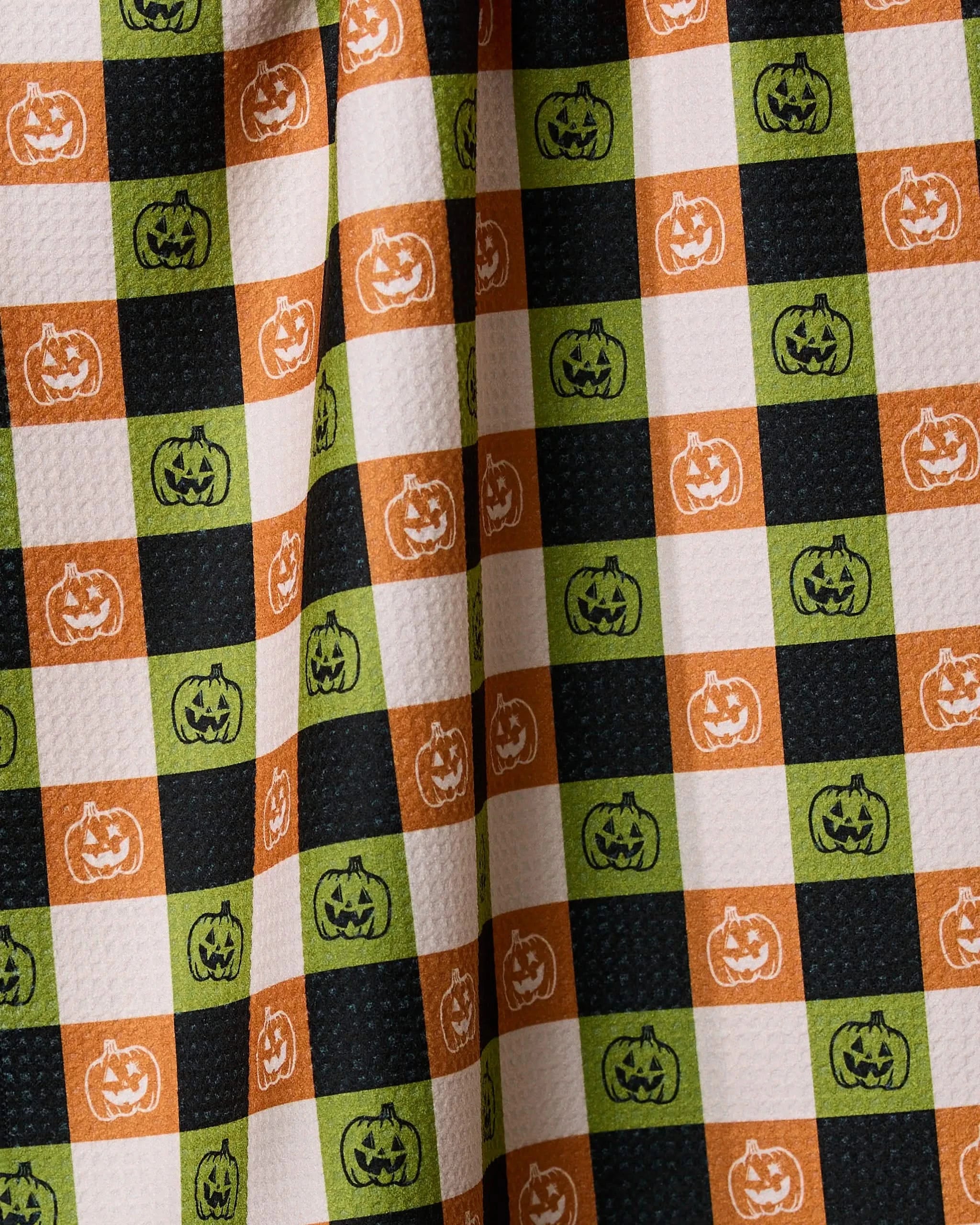 The Geometry Jack o Lantern Checker tea towel features orange, green, black, and white checkered squares with smiling jack-o-lanterns for a festive Halloween touch—crafted from recycled materials.