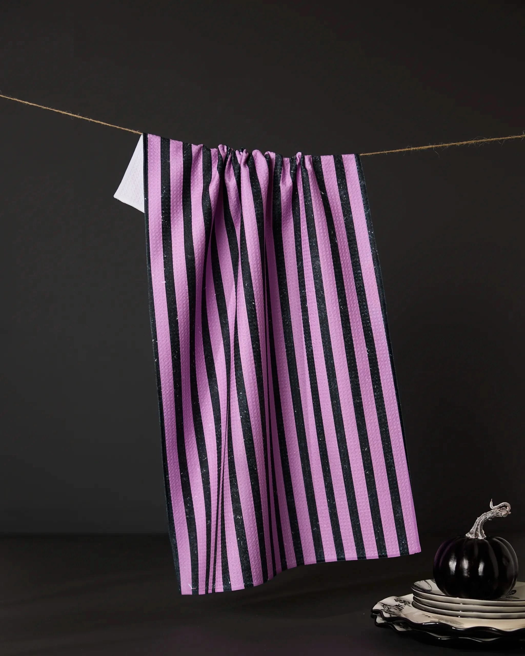 The Neon Halloween Nights tea towel by Geometry, featuring purple and black vertical stripes and made from recycled materials, hangs on a string against a dark backdrop near white plates and a small dark onion.