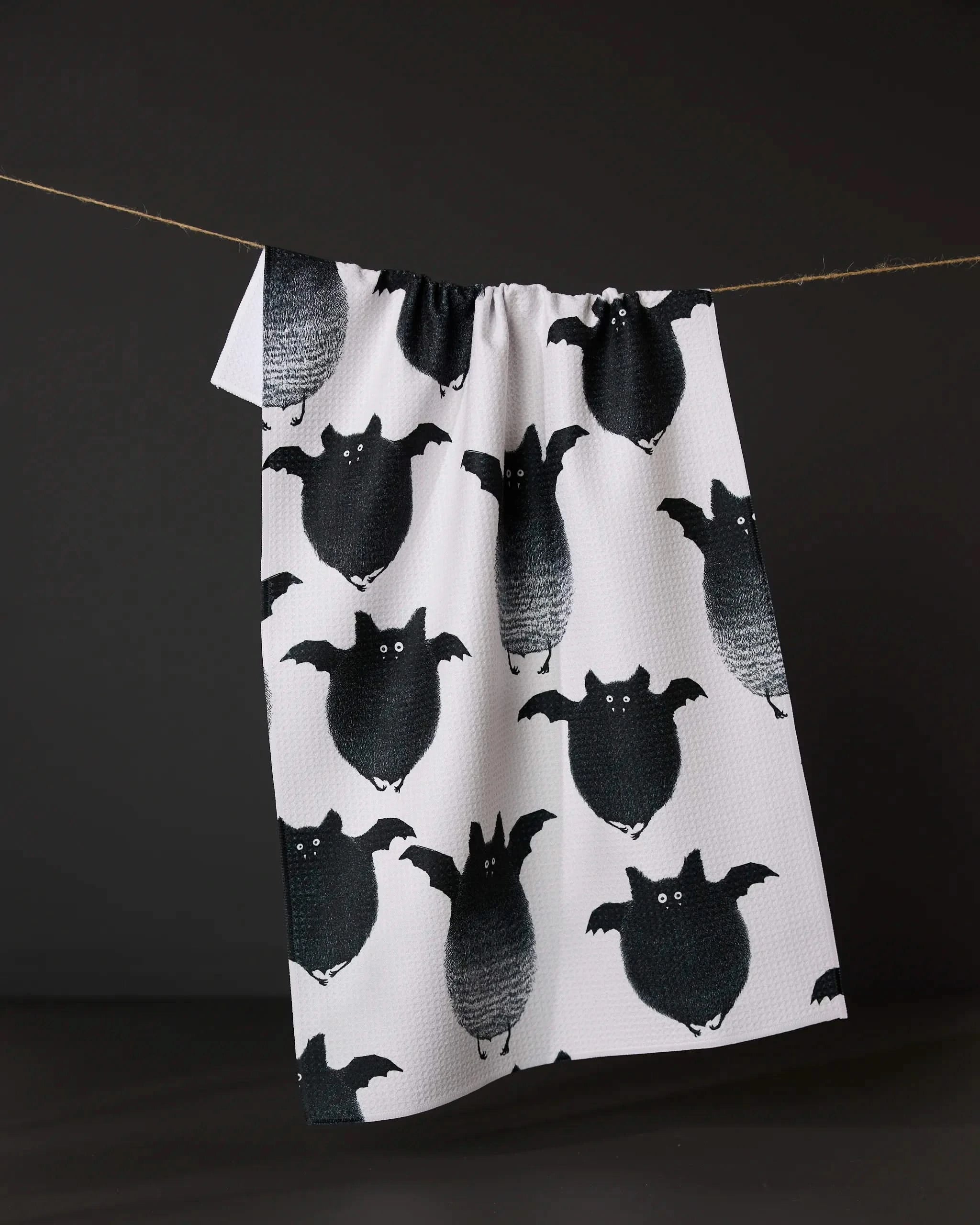 The Geometry Fuzzy Bats tea towel, featuring a pattern of cute round black bats, hangs from a string against a dark background.