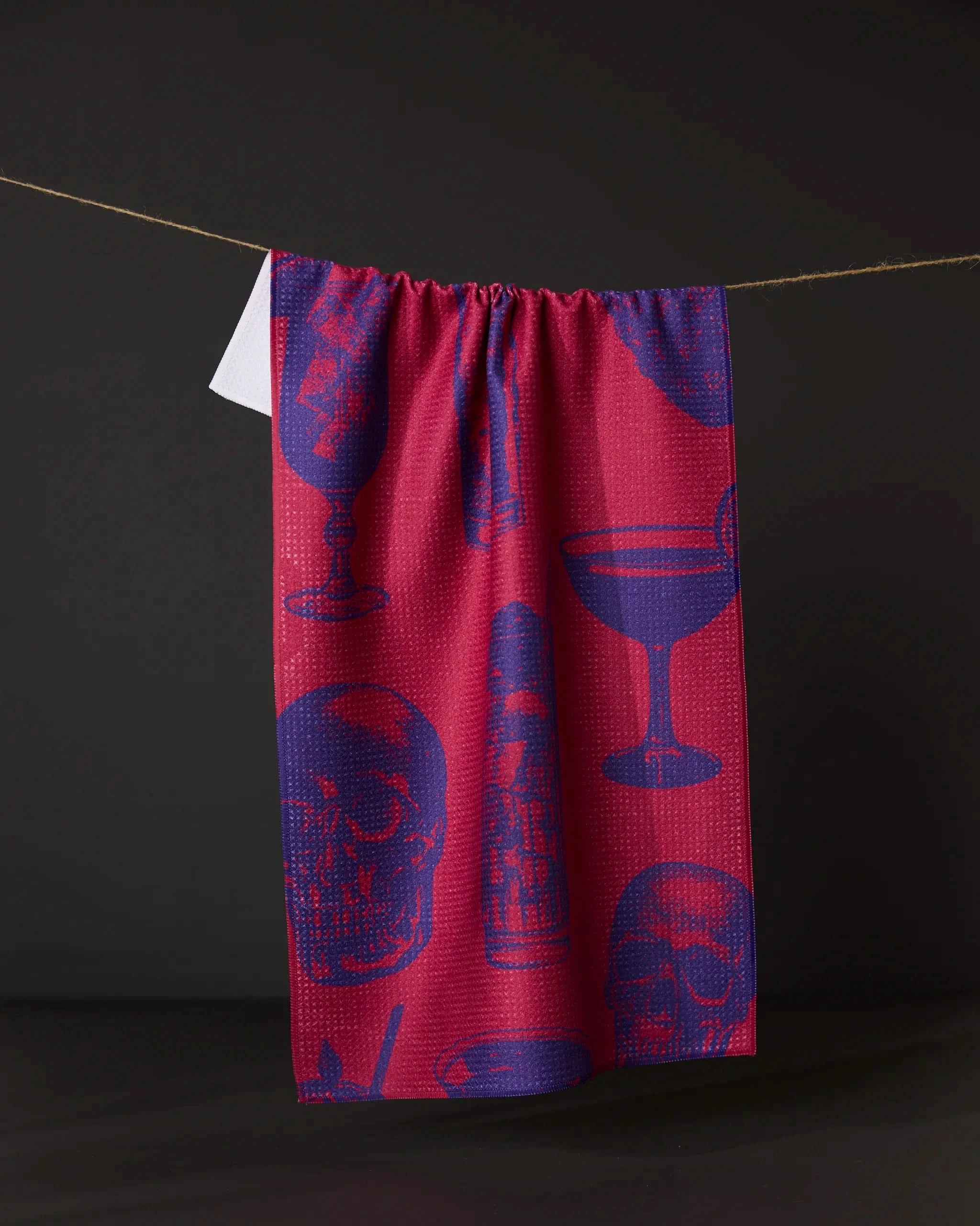 The Geometry Ghostly Cheers Red eco-friendly kitchen towel, featuring blue skull and cocktail glass patterns, hangs on a string against a dark background.