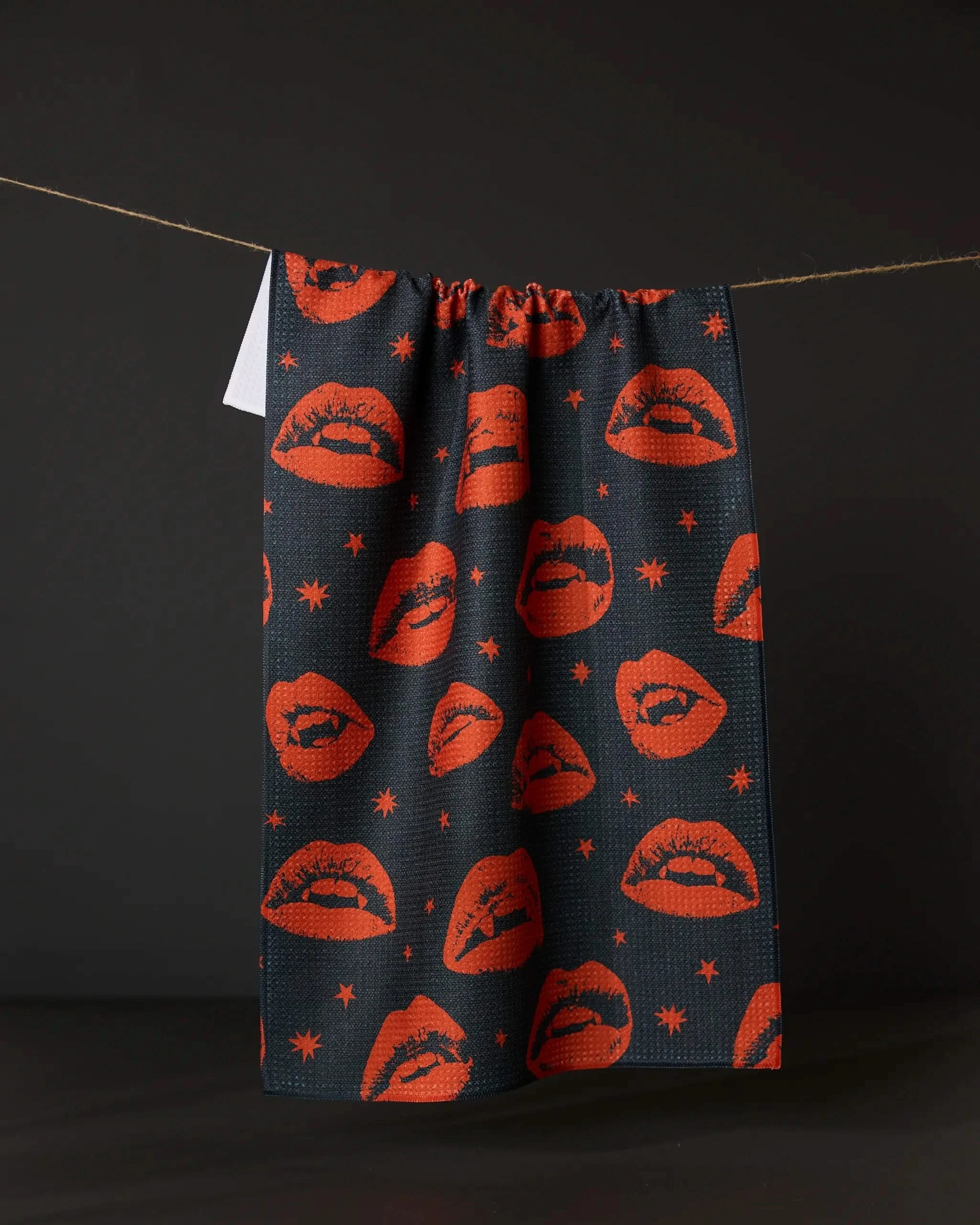 The Geometry Fangs tea towel, made from recycled materials, features a pattern of red lips and small stars and hangs from a string against a solid black background.