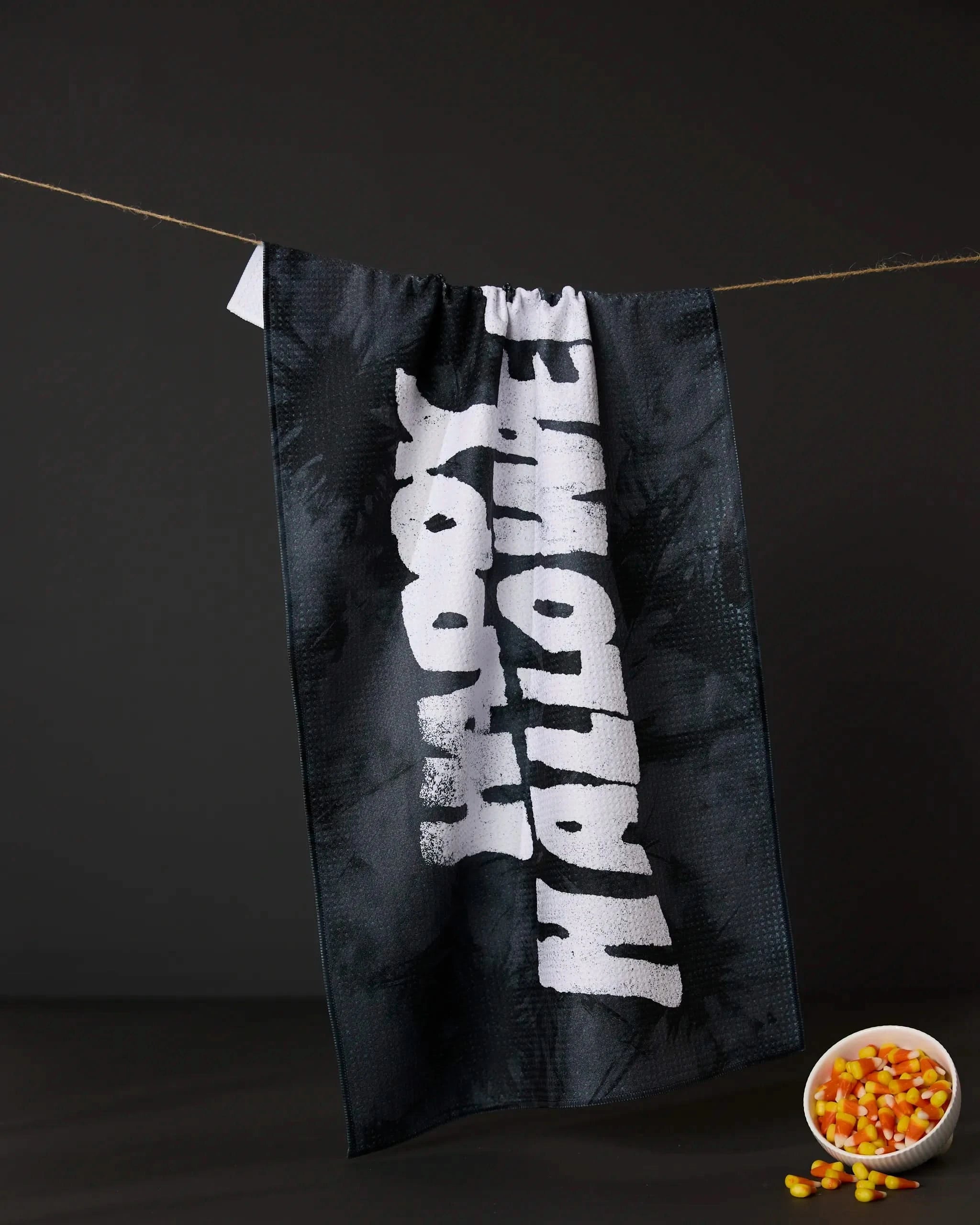 The Geometry Happy Halloween Night tea towel, in black with bold white lettering, hangs on a clothesline against a dark backdrop. A small white bowl of candy corn appears in the bottom right corner.