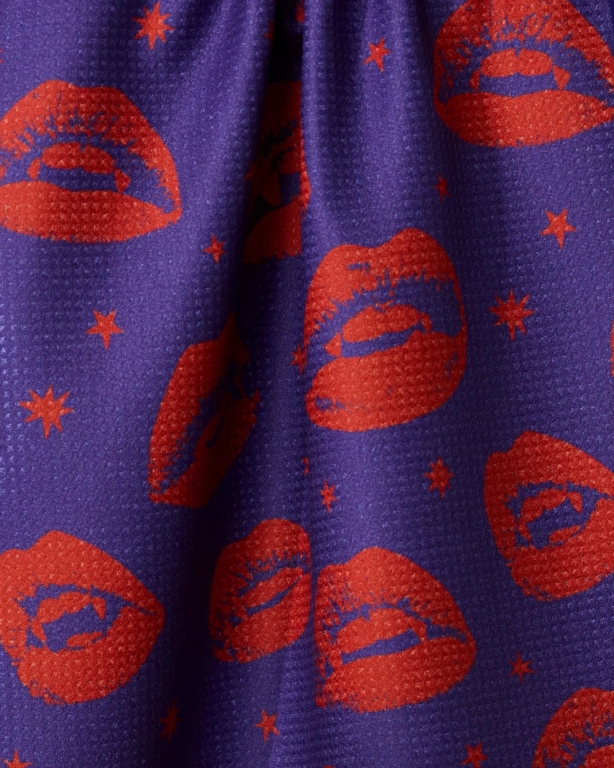 The Geometry Fangs Purple tea towel features red lipstick lip prints and small red stars on purple, made from recycled materials with a textured finish. Gathered in the center, it creates soft folds and gentle shadows.
