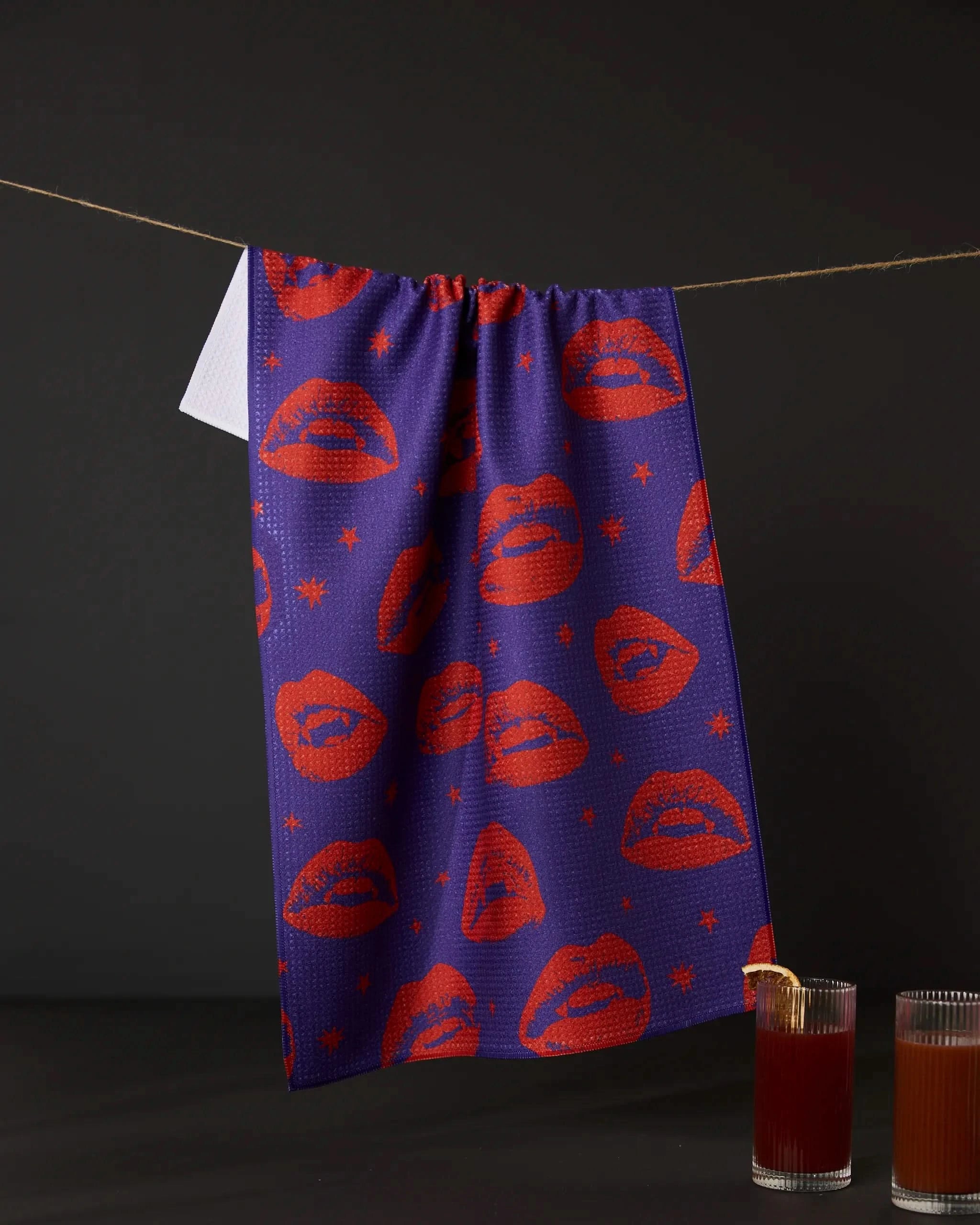 The Geometry Fangs Purple tea towel with red lips and stars, made from recycled materials, hangs on a string against a dark background, with two glasses of red liquid below on the right.
