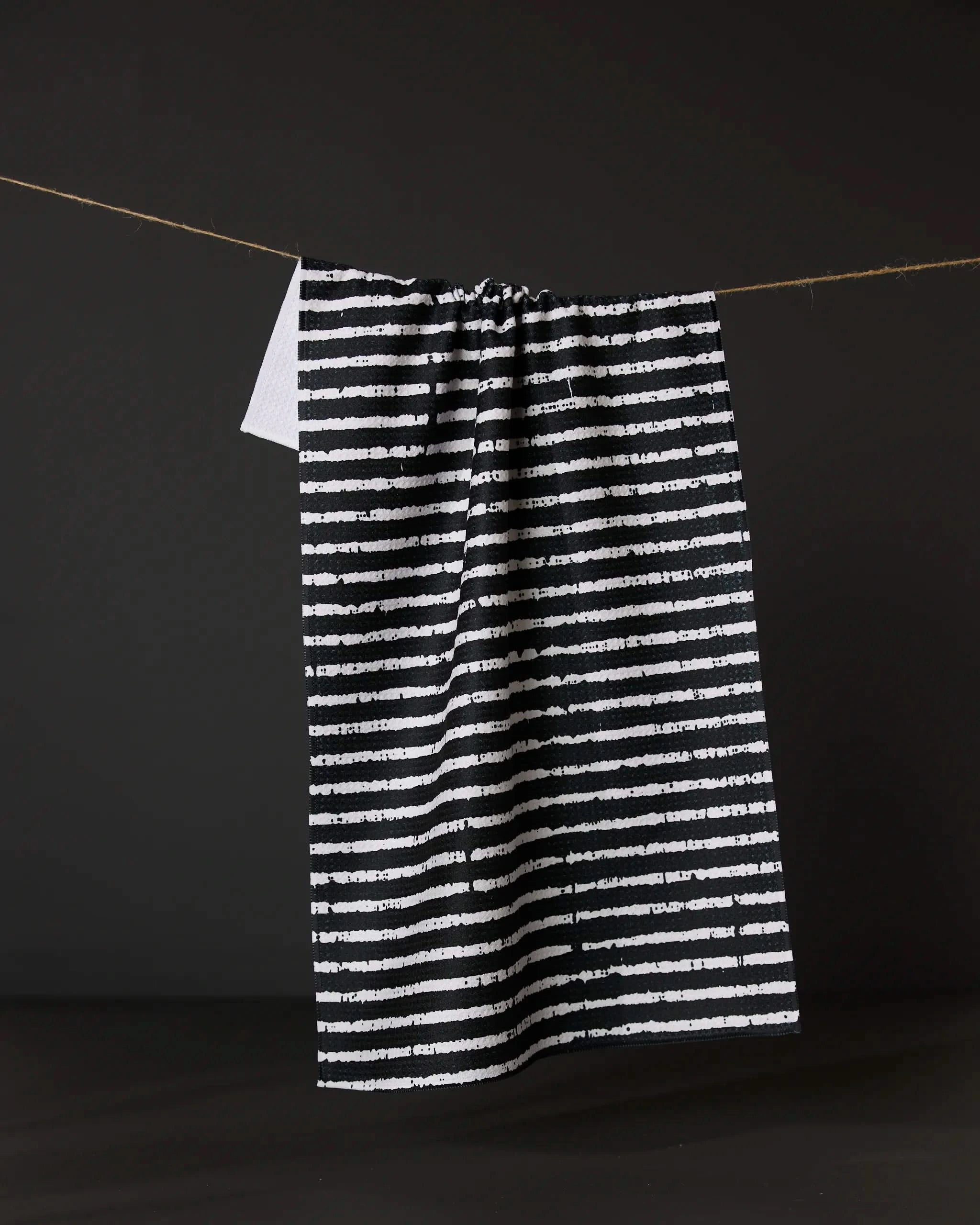 The Eerie Stripe tea towel by Geometry, featuring black and white stripes and a small white tag at the top, hangs on a string against a dark background.