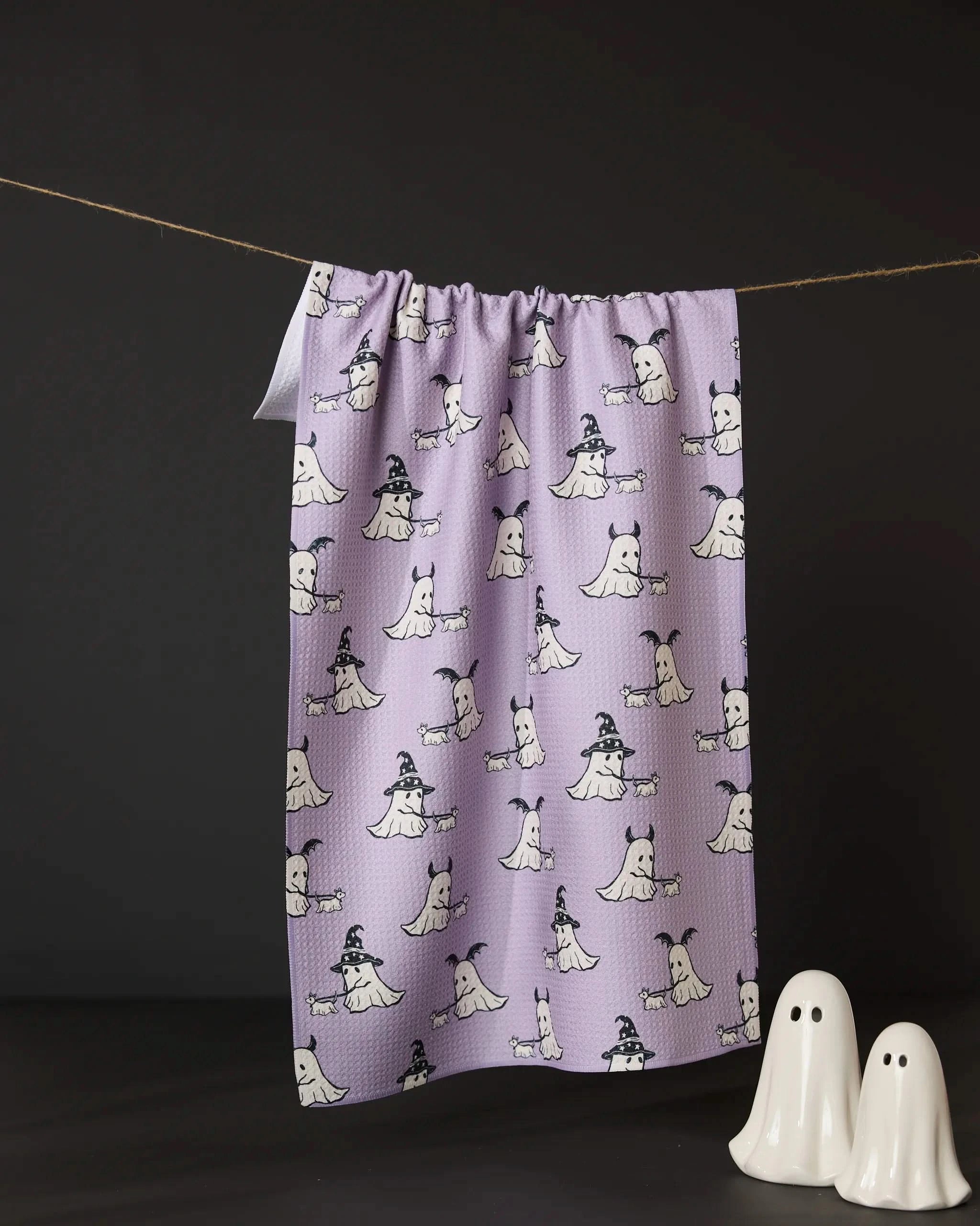 The Geometry Spooky Ghost Walk tea towel, featuring cartoon ghosts—some in witch hats—hangs on a string against a black background, with two white ghost figurines nearby. Made from recycled materials for eco-friendly charm.