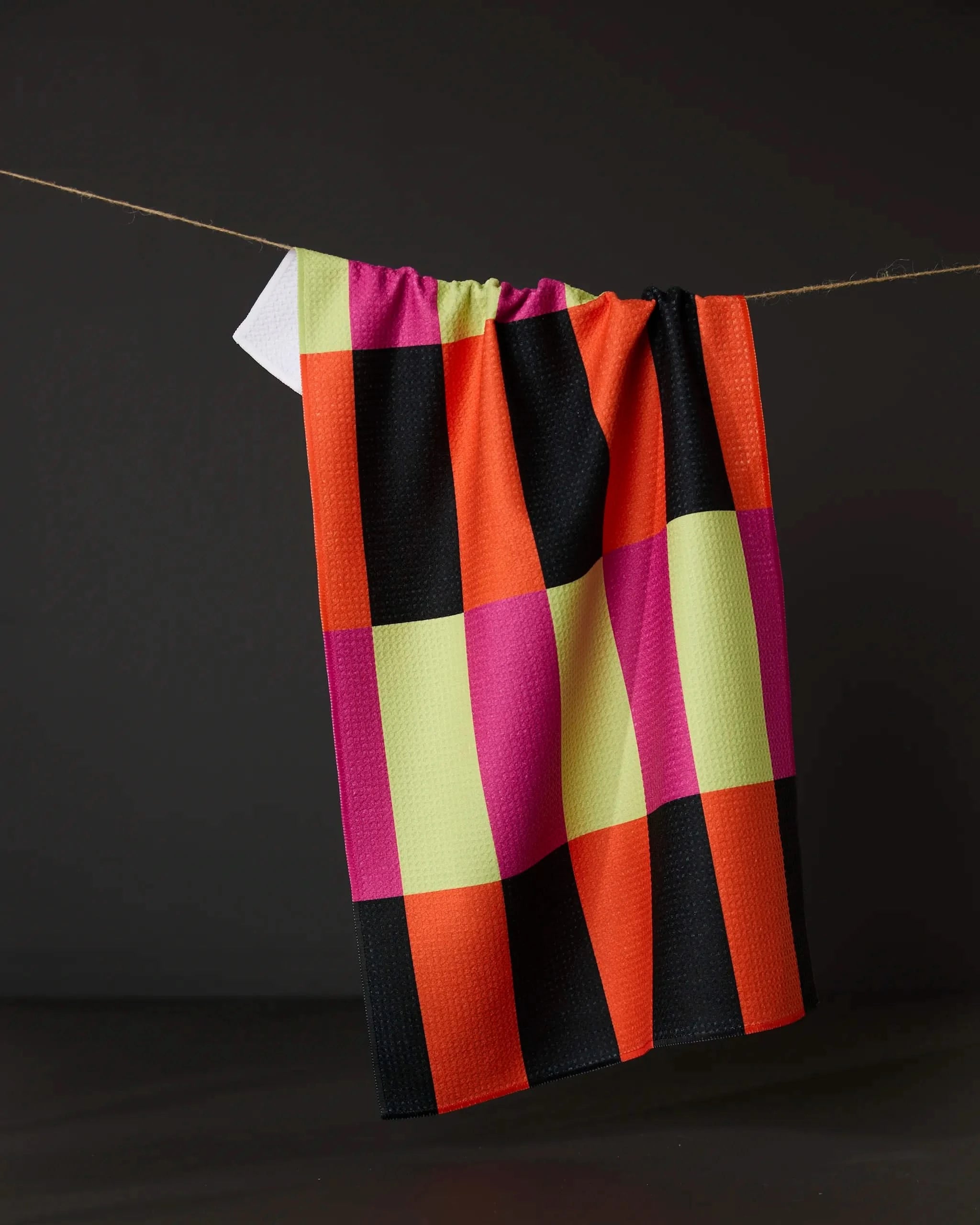 The Geometry Spooky Hour Stripe tea towel, featuring bold black, orange, red, magenta, and yellow checks, hangs against a dark background. Made from ultra-absorbent recycled materials for a stylish and sustainable kitchen.