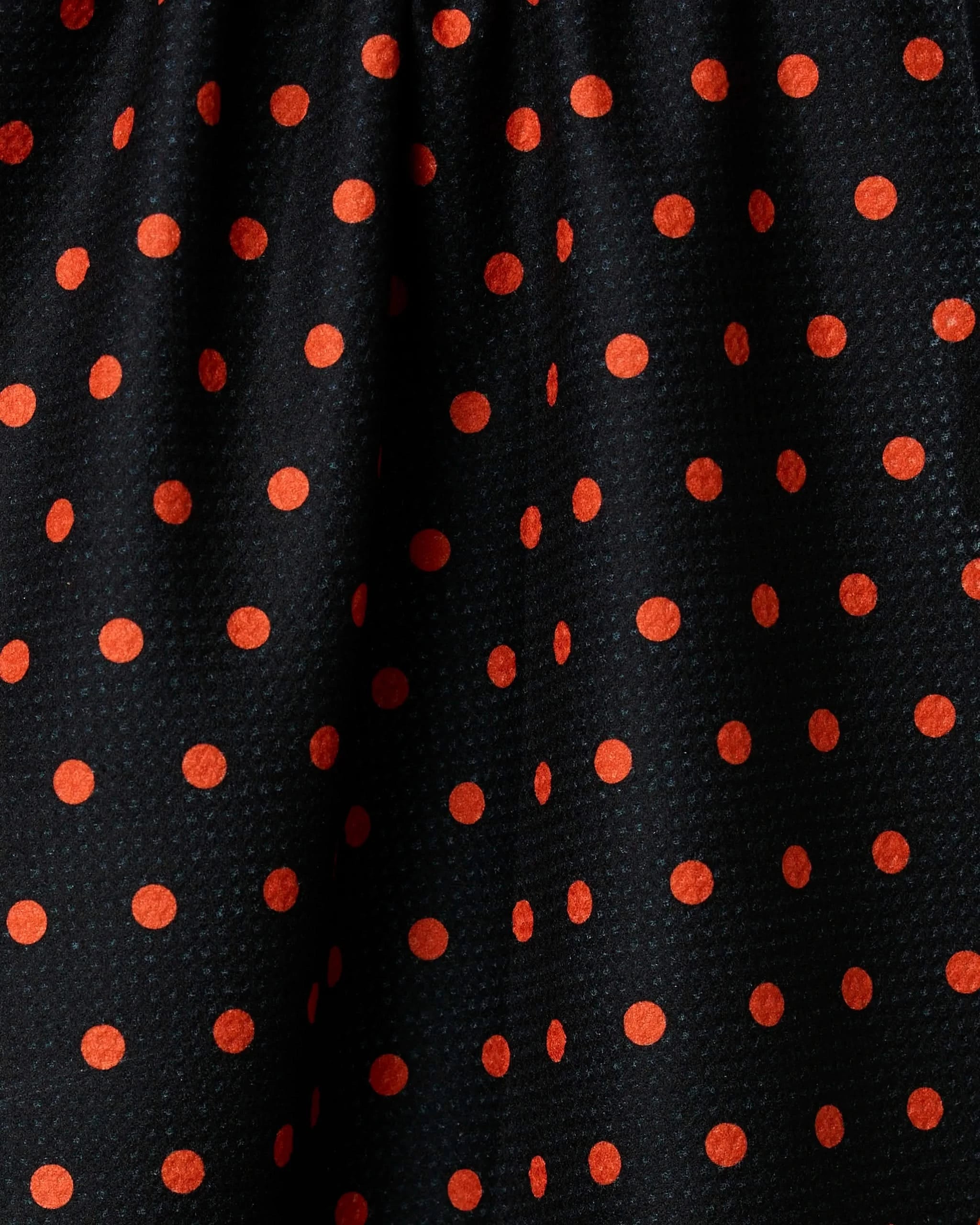 Spottie by Geometry features black fabric with evenly spaced orange polka dots. Made from recycled materials, it has a slight texture and is softly gathered at the top for elegant folds and shadows.