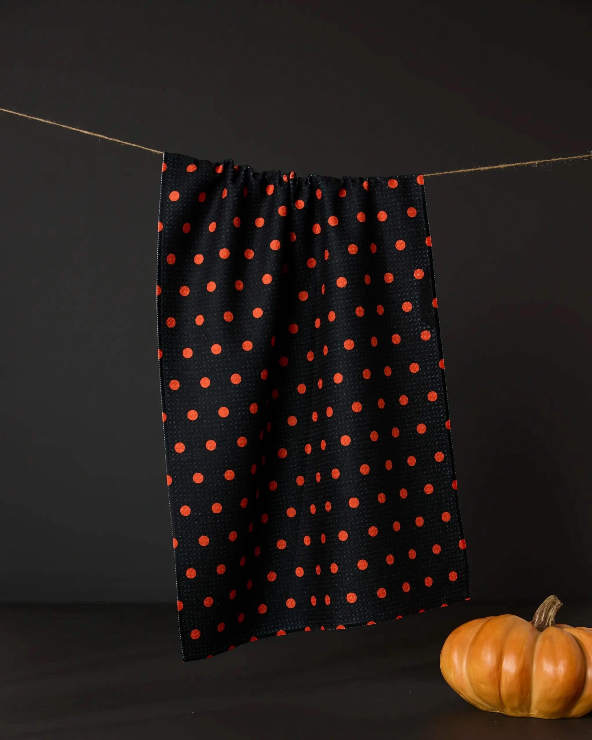 The Geometry Spottie tea towel features an orange polka dot pattern and hangs against a dark backdrop with a small pumpkin below. This absorbent kitchen towel combines style and practicality for any kitchen.