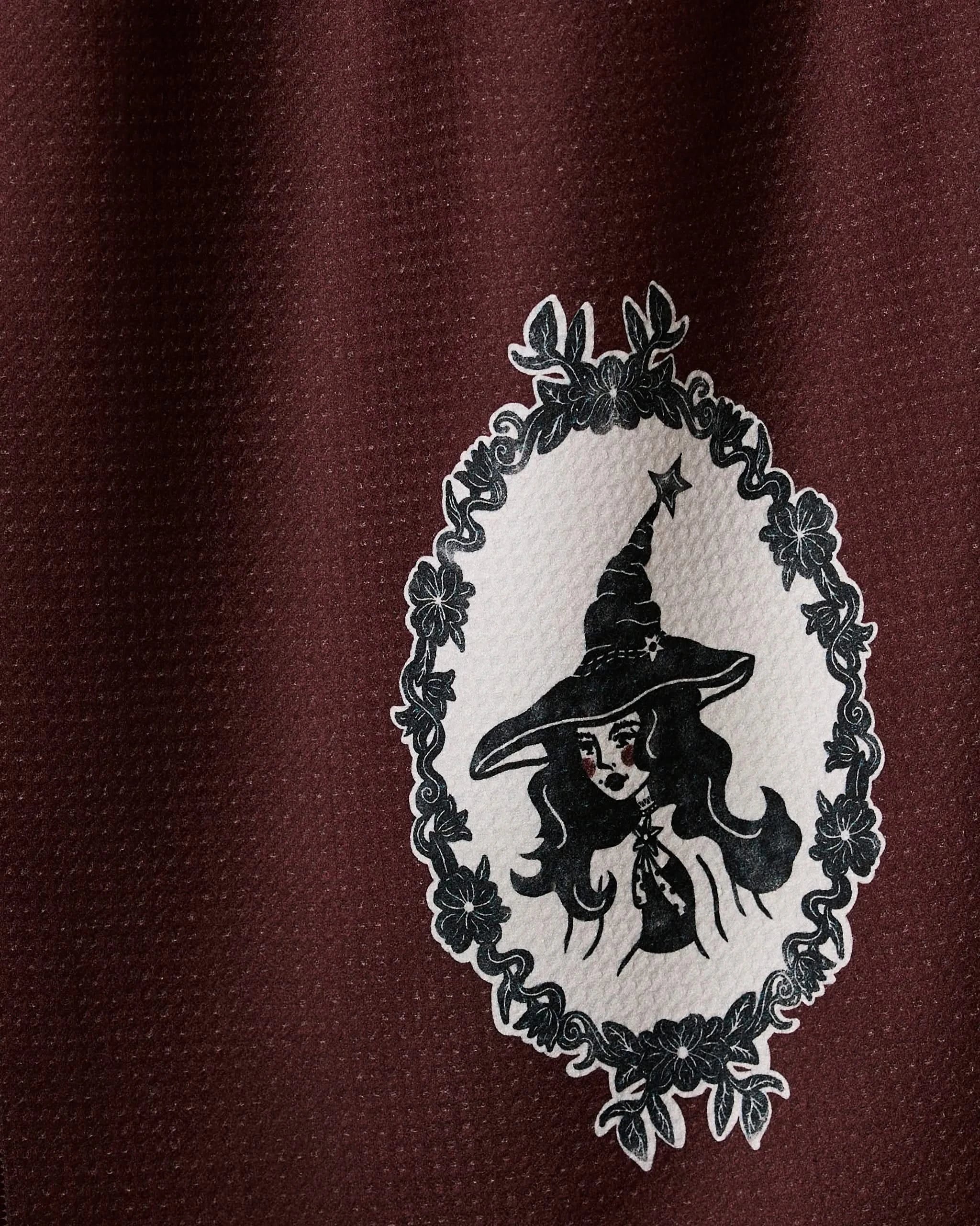 “Portrait of a Cool Witch” by Geometry features a vintage-style witch in an ornate oval frame, printed on quick-drying dark red fabric—a unique tea towel for any kitchen.