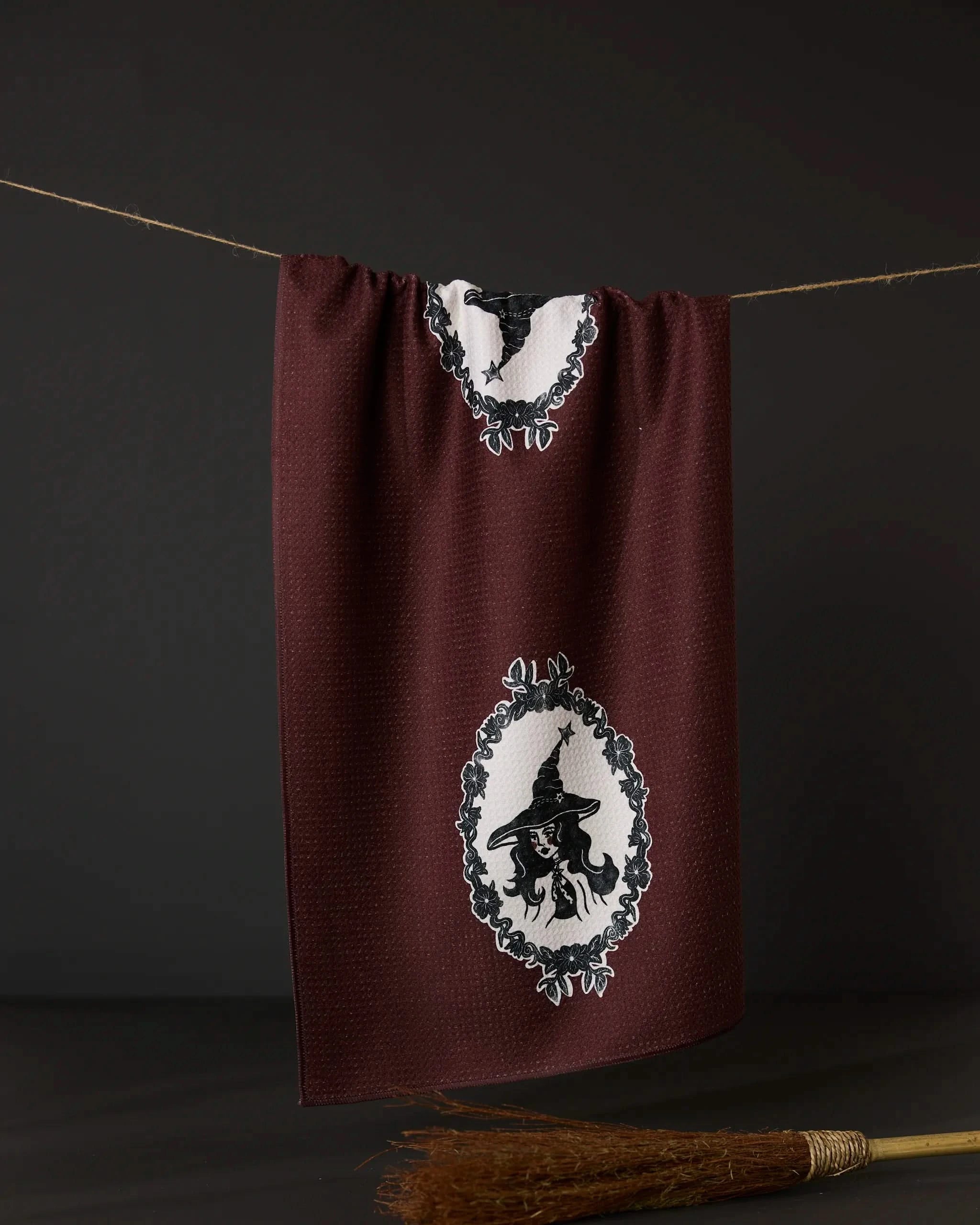 The Geometry Portrait of a Cool Witch tea towel features black and white framed silhouettes of a hatted witch, with a broom below, against maroon. Crafted from recycled materials, it hangs elegantly against a dark background.