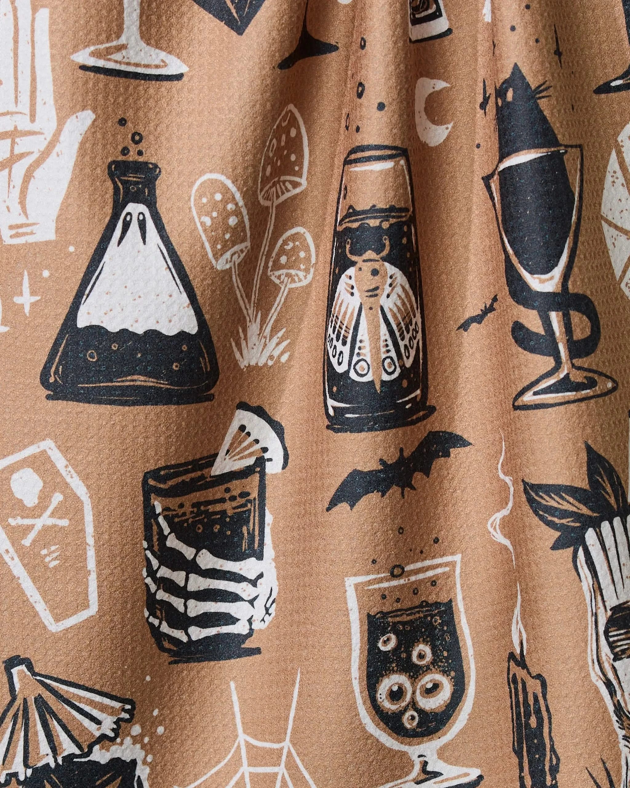 The Spooky Hour towel by Geometry is brown and ultra-absorbent, featuring black and white spooky illustrations like a bubbling flask, mushrooms, moth in a jar, eyeball cocktail, bat, coffin, spiderwebs, and tarot cards.