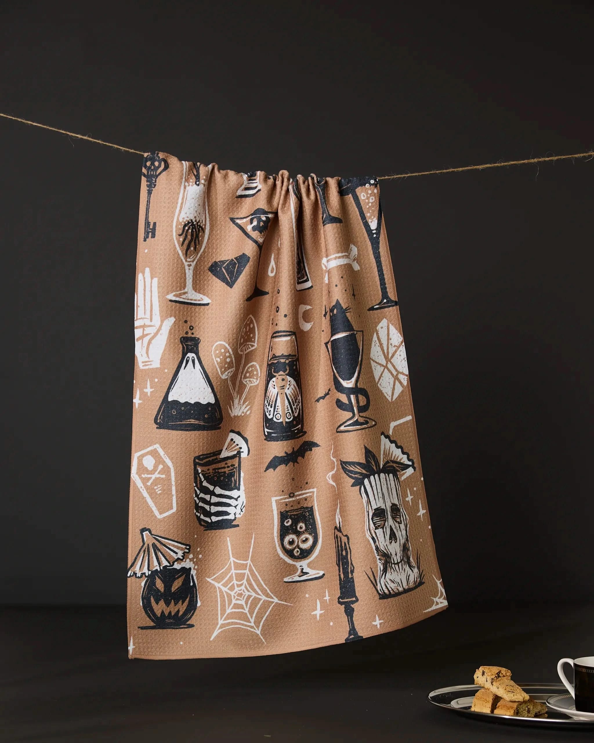The Spooky Hour tea towel by Geometry—featuring white and black jack-o’-lanterns, potions, candles, a skull, and spiderwebs—hangs on a string. Nearby, a plate with biscotti and coffee rests on a dark surface.