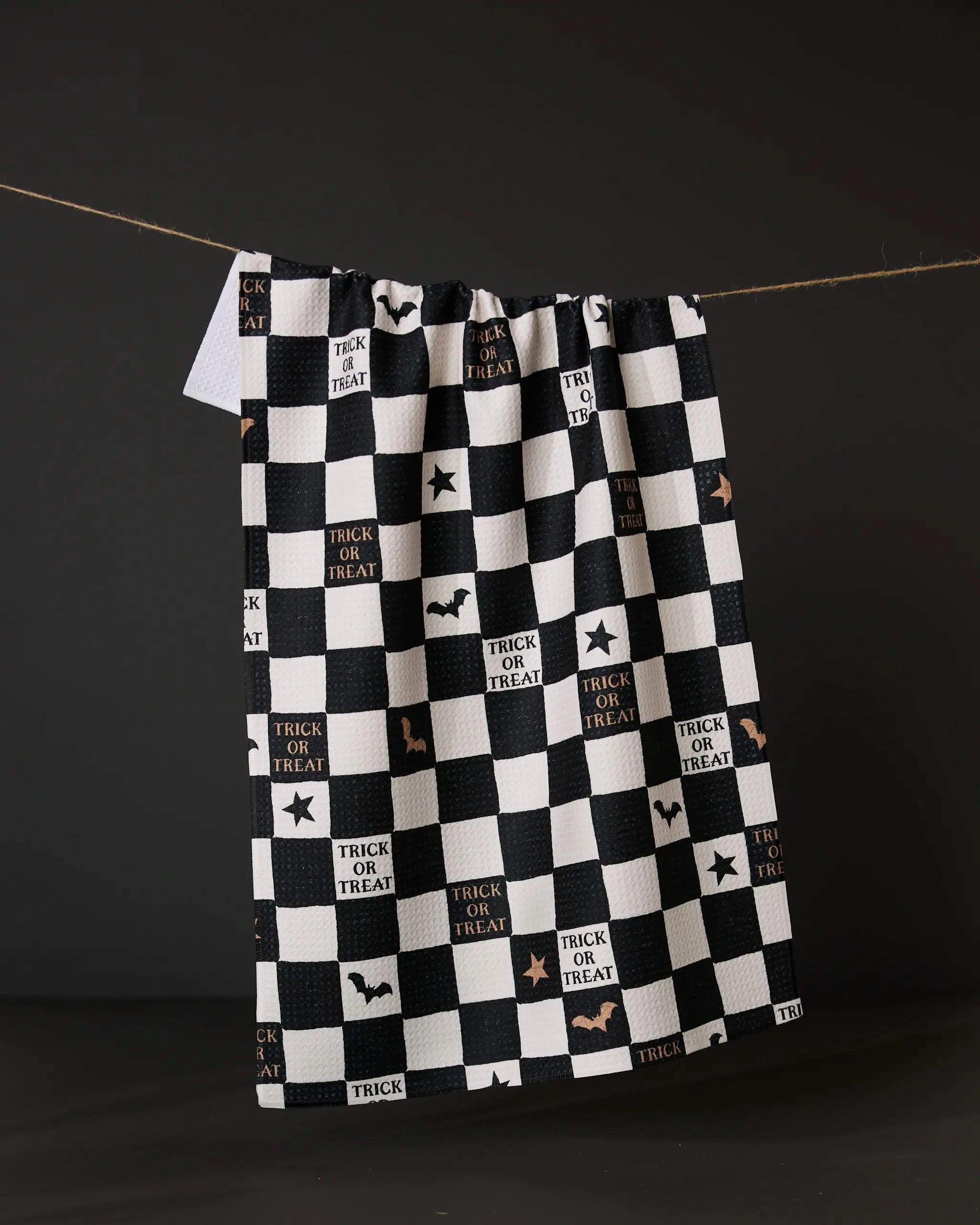 The Tricks and Treats kitchen towel by Geometry features black and white checkers, eco-friendly fabric, Halloween designs with bats, stars, and Trick or Treat—all displayed hanging against a dark background.