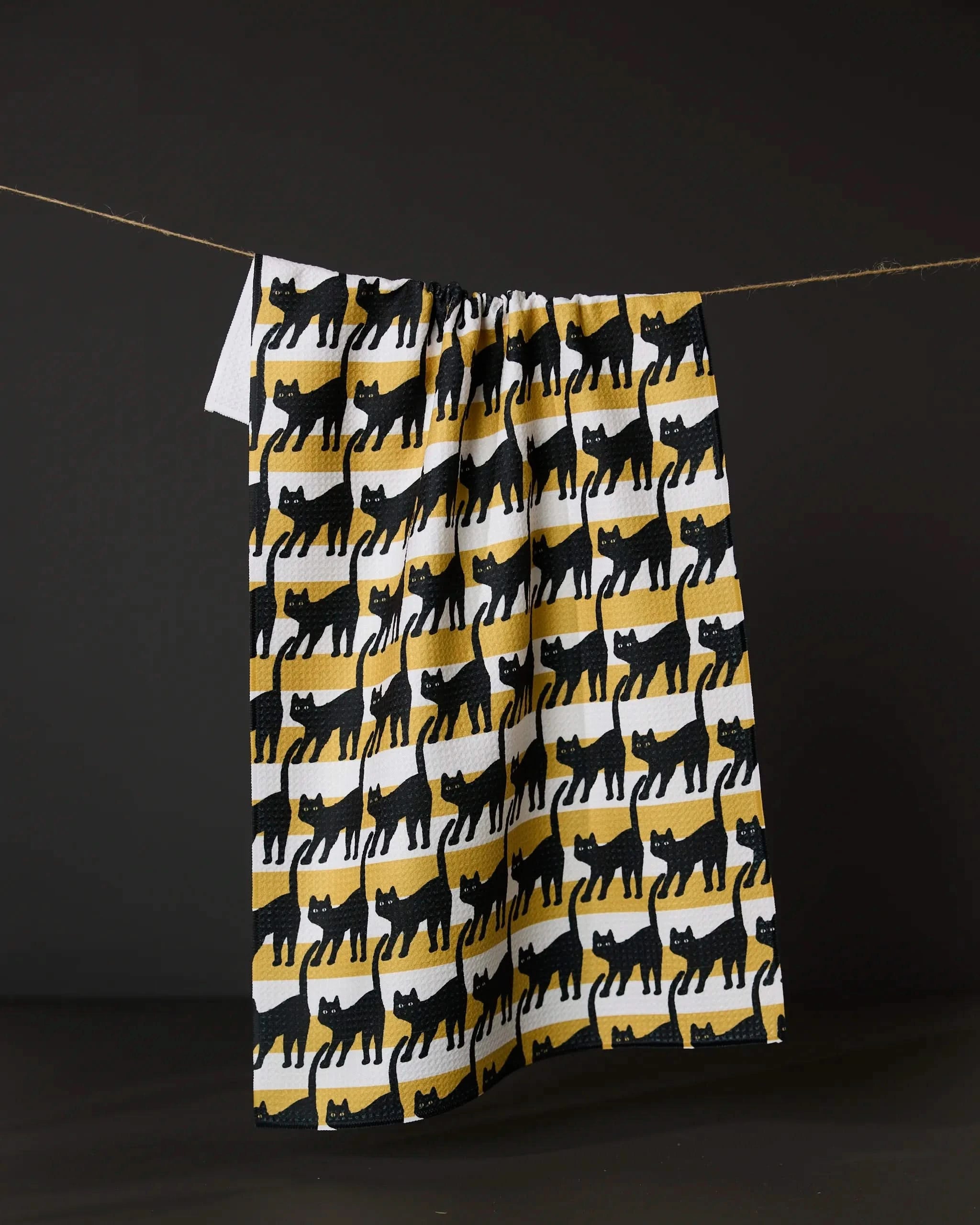 The Geometry Salem Cat Stripe kitchen towel, made from eco-friendly materials, showcases black cat silhouettes on white and yellow striped patterns, hanging on a string against a dark background.