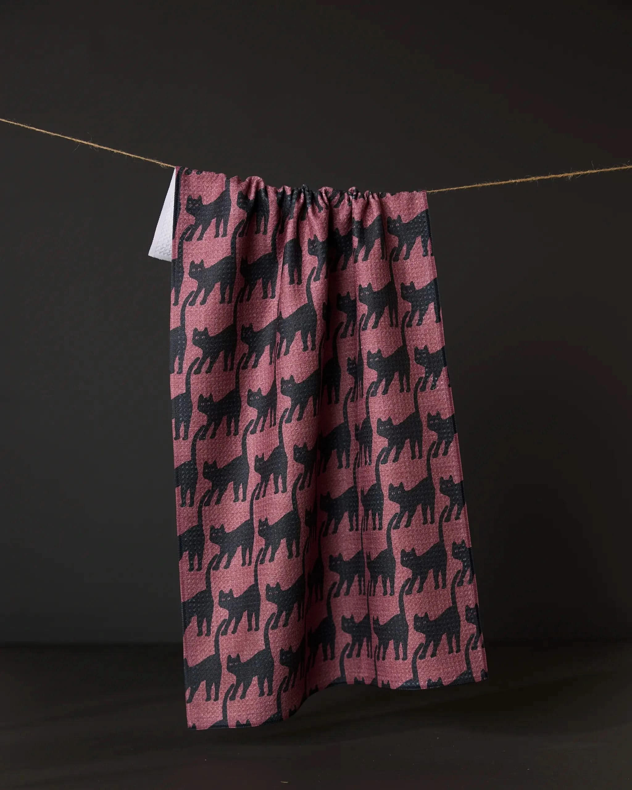 The Geometry Salem Cat tea towel, made from recycled materials, showcases a repeating black cat silhouette pattern on a maroon background and hangs against a dark backdrop.