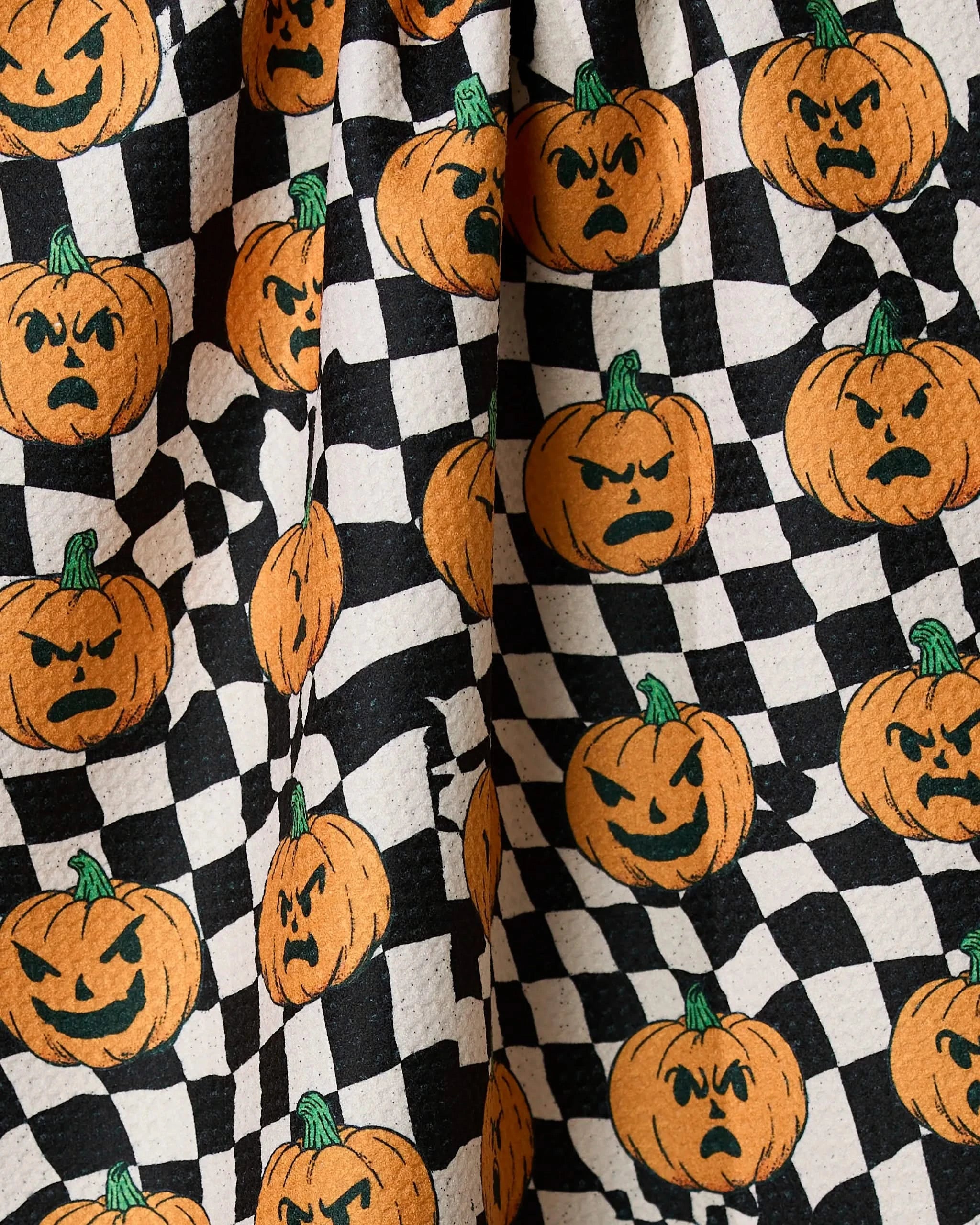 Close-up of Geometrys Sinister Jack Os recycled kitchen towel, featuring a black-and-white checkered pattern with orange pumpkins showing angry or mischievous faces and green stems.