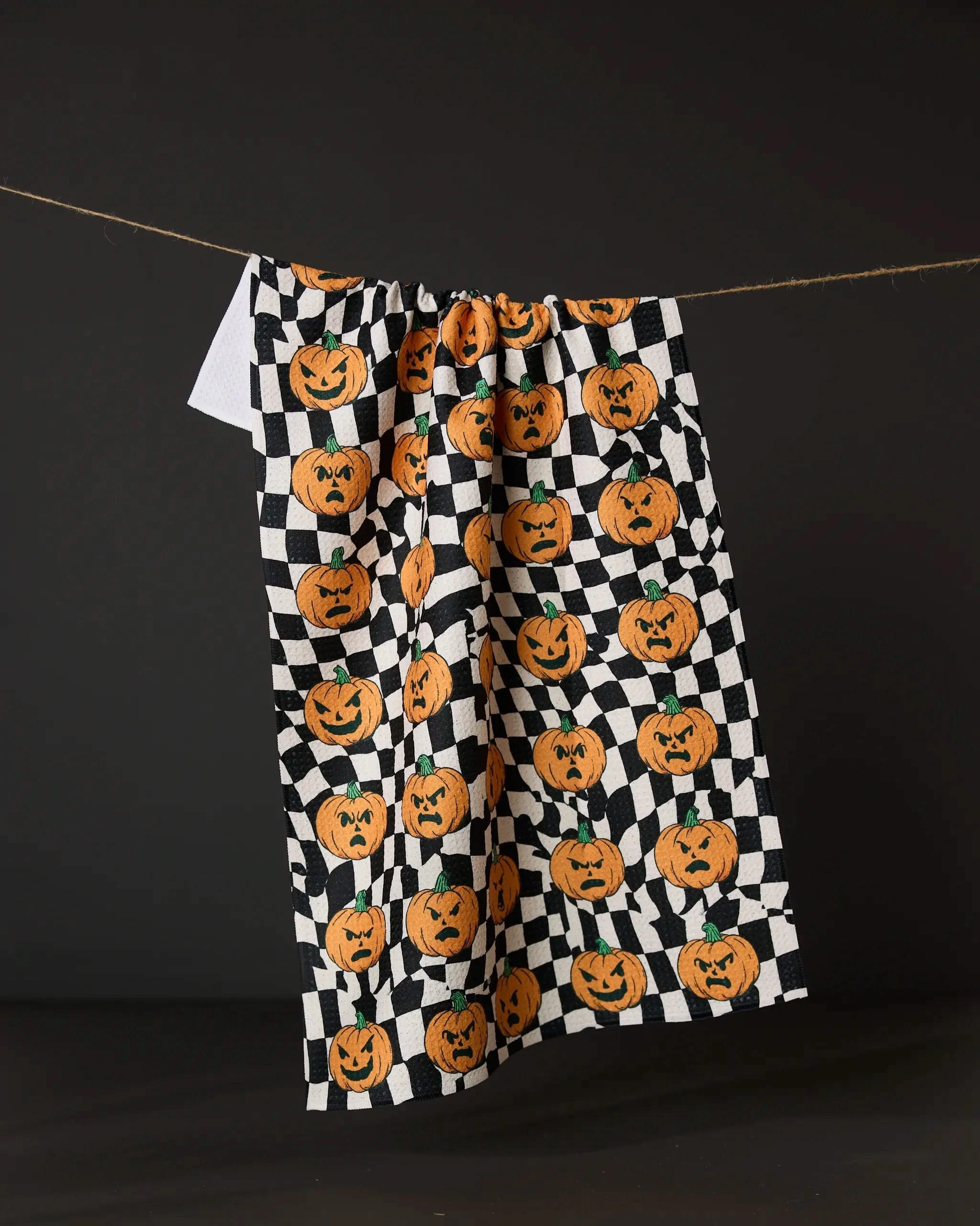 The Geometry Sinister Jack Os tea towel features a black and white checkered pattern with orange jack-o-lantern pumpkins, hanging against a dark backdrop.