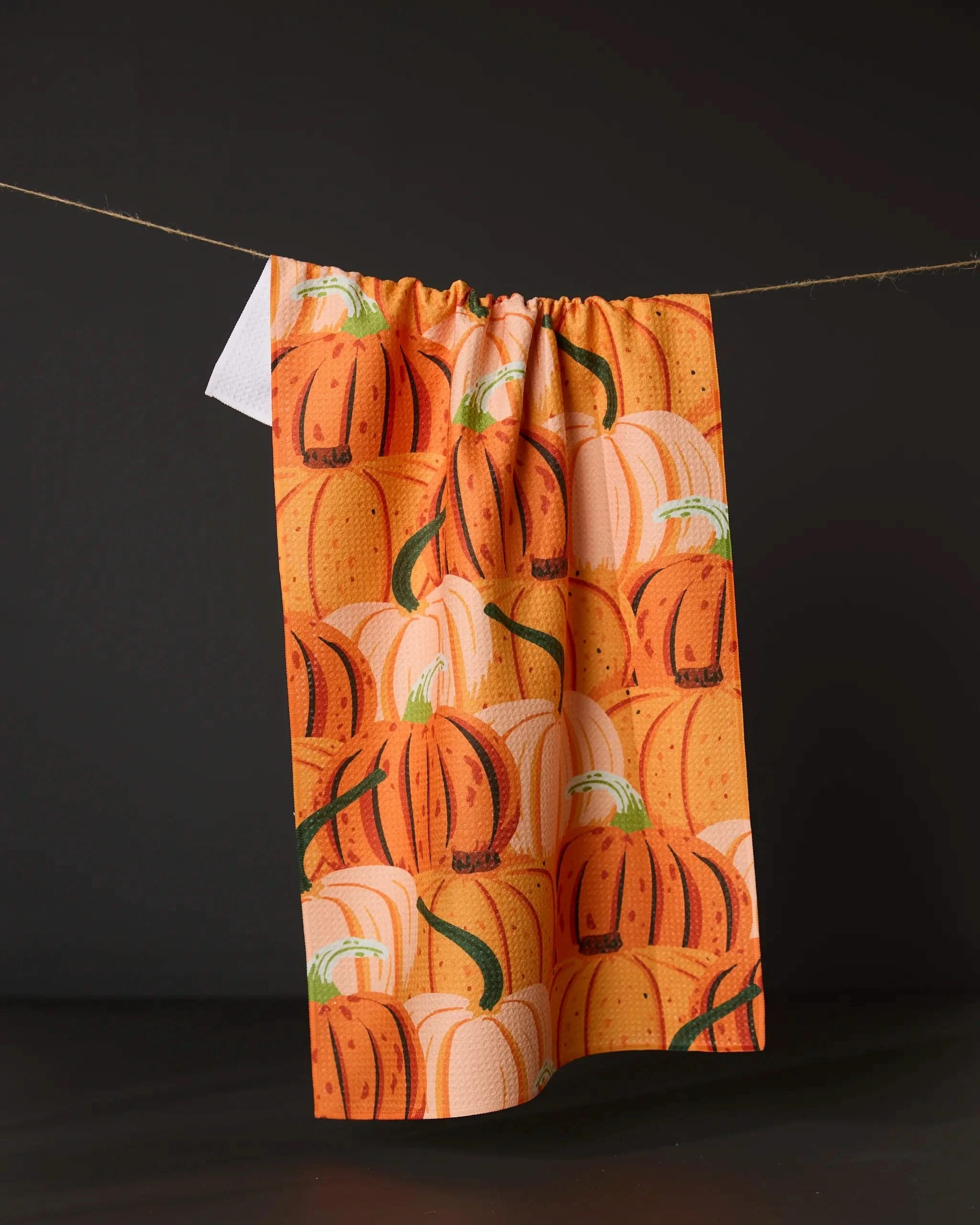 The Geometry Rows of Gourds tea towel, featuring an ultra-absorbent design and an orange and white pumpkin pattern, hangs on a string against a dark background.
