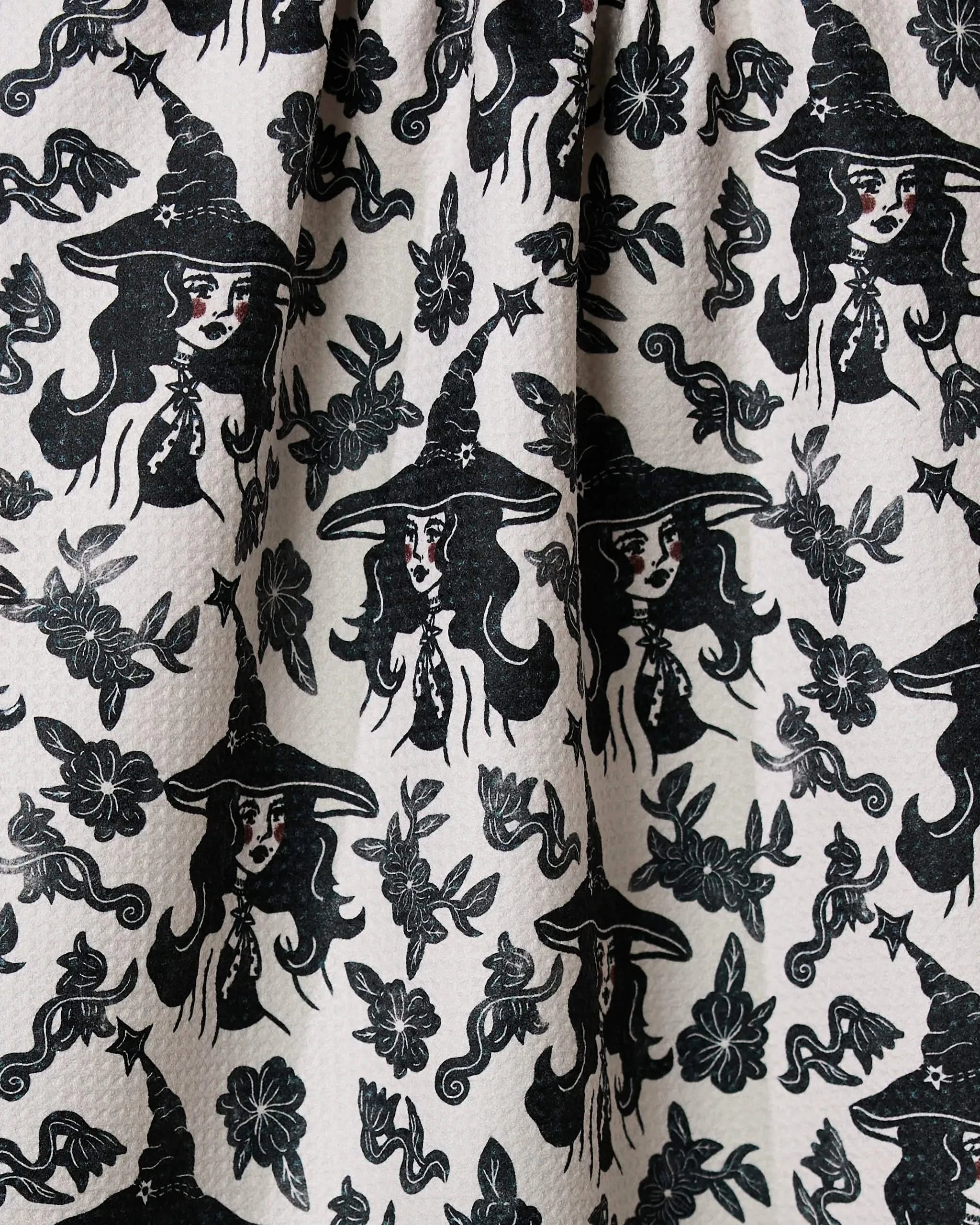 The Geometry Floral Witches tea towel showcases a vintage black and white hand-drawn design with women in witch hats, snakes, stars, flowers, and moons. Crafted from ultra-absorbent recycled materials for style and sustainability.