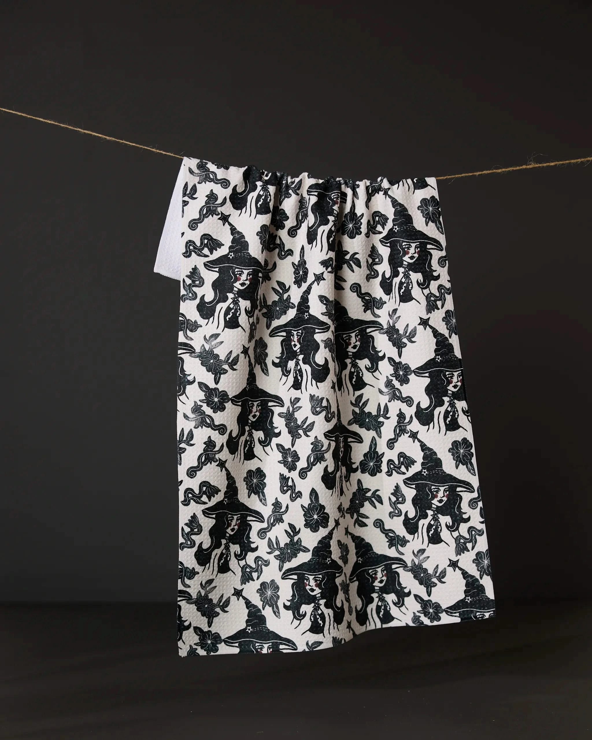 The Floral Witches tea towel by Geometry, made from recycled materials, showcases black tiger and floral patterns on white fabric and hangs on a string against a dark backdrop.
