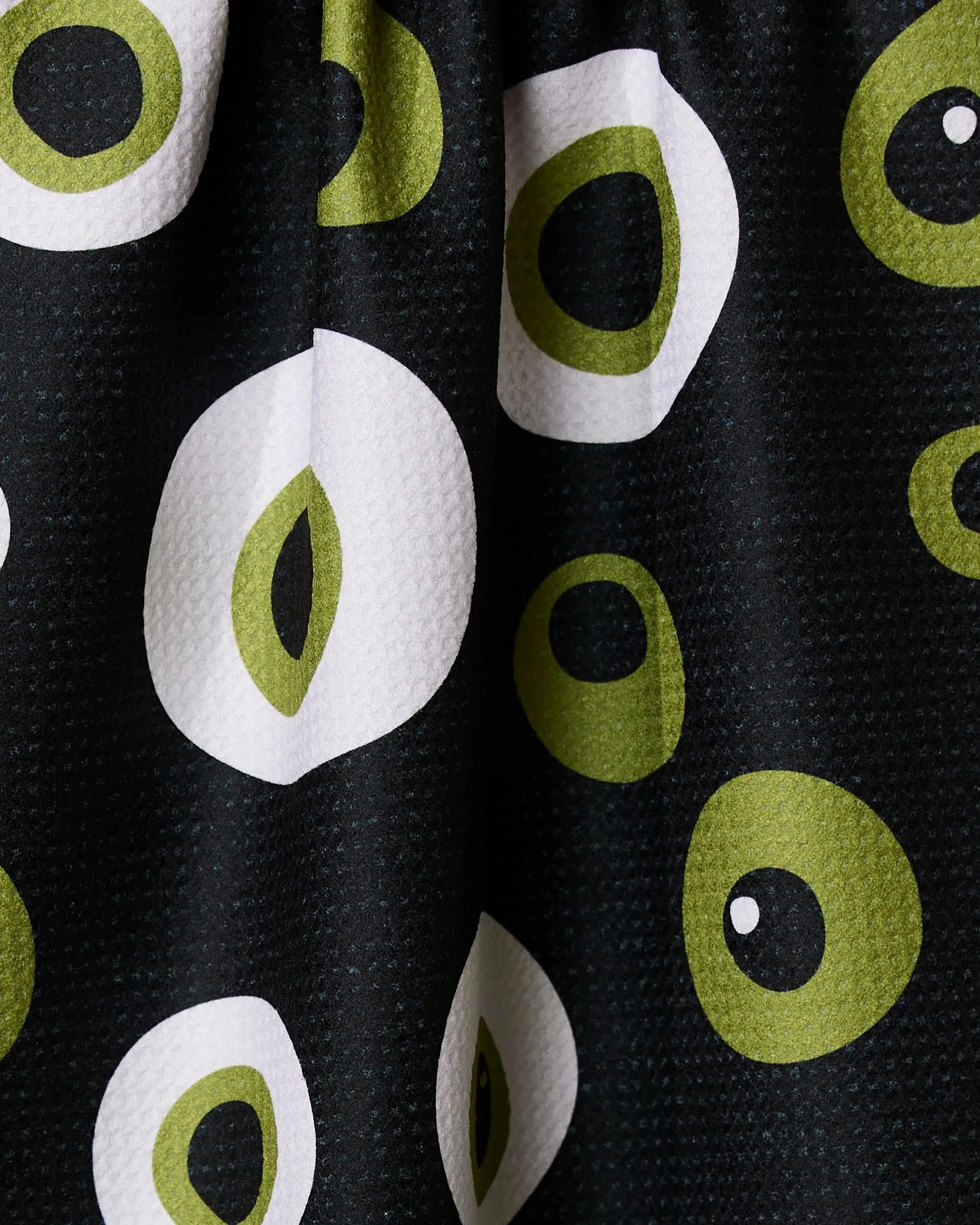A close-up of Geometrys Tricky Eyes tea towel, made from recycled materials, shows black ultra-absorbent fabric with green and white abstract circular and oval patterns repeated in varying sizes and gentle folds.