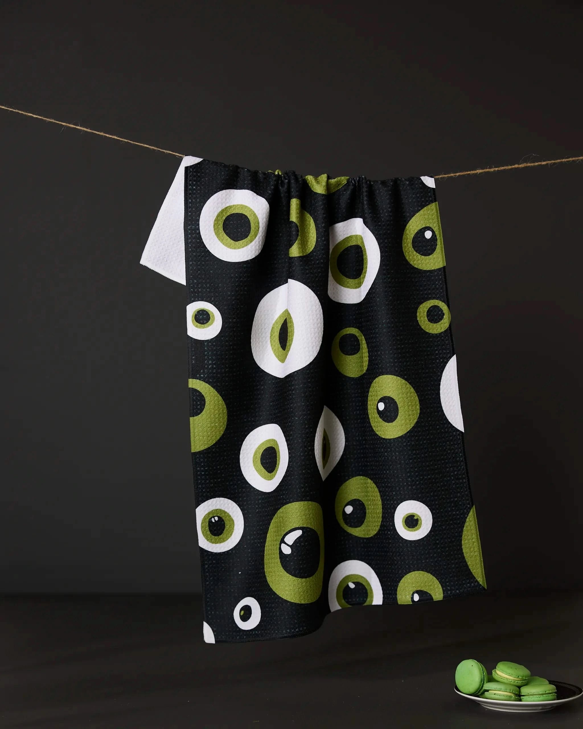 The Tricky Eyes tea towel by Geometry, featuring green and white circular patterns on black, hangs on a clothesline against a dark backdrop with green macarons on a plate below.
