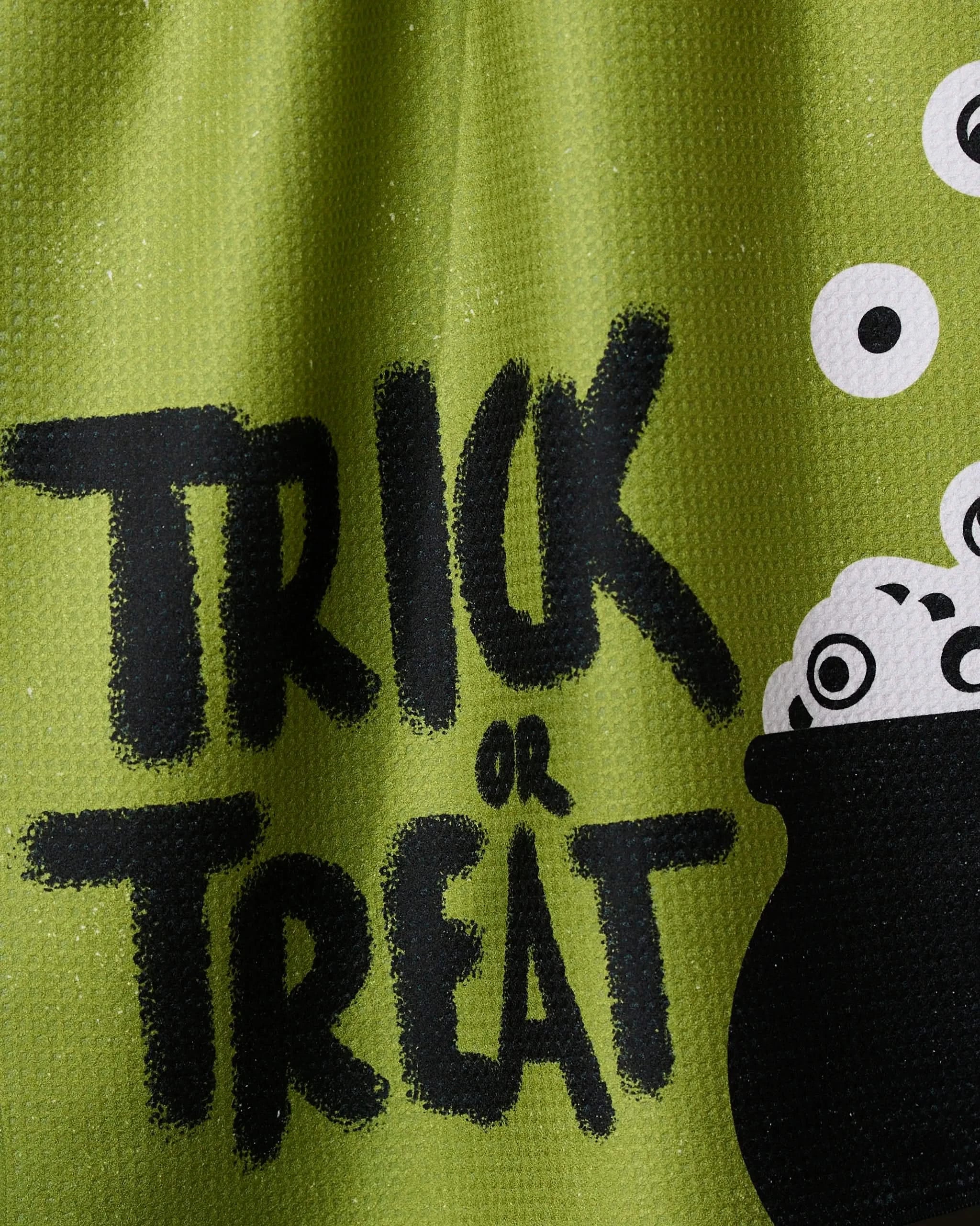 A close-up of Geometrys Tricky Treats green tea towel, featuring bold black Trick or Treat text beside a black cauldron filled with white eyeballs.
